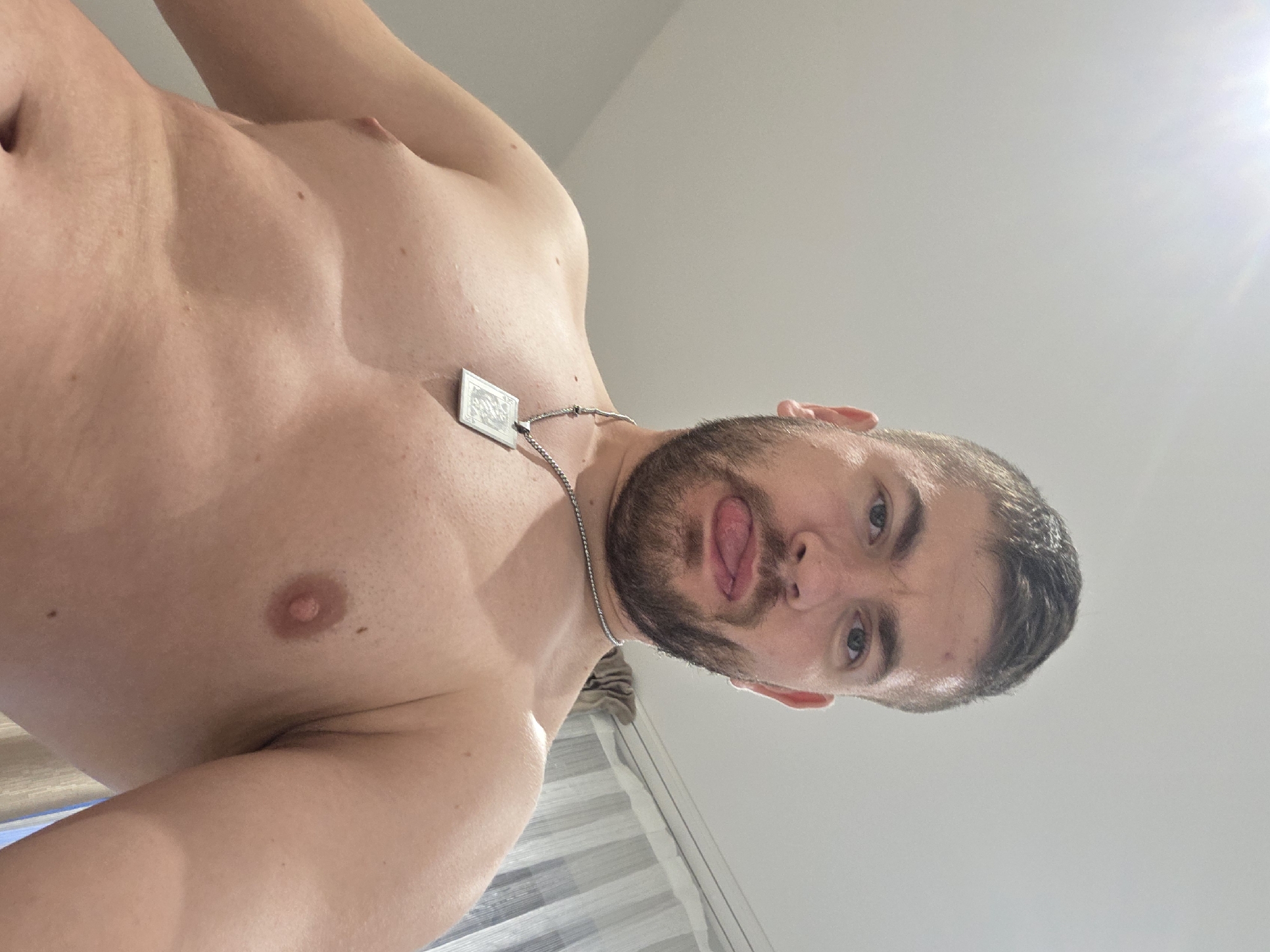 A shirtless man with a beard and a silver necklace is looking down at the camera and sticking his tongue out. He is indoors with a white ceiling above and a white curtain behind him. The location in Yagodovo, Bulgaria is not discernable from the image.