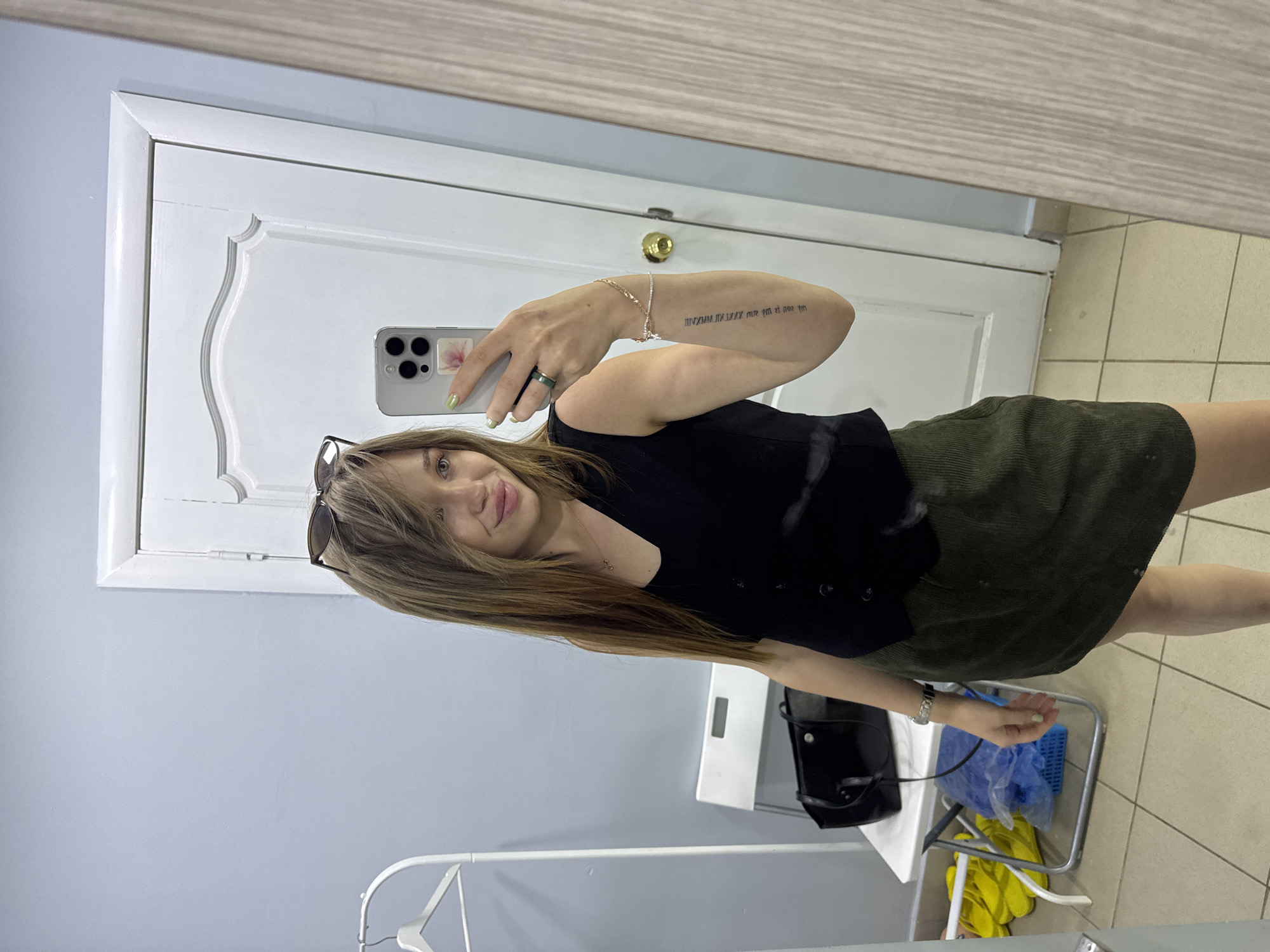 A young woman with long, brown hair and light skin is taking a selfie in a room with light blue walls and a white door. She is wearing a black vest over a black v-neck top and a dark olive-green corduroy skirt. Her right arm is extended, holding a smartphone with a pink flower sticker on the back. She is wearing a green ring on her left index finger and a bracelet on her right wrist. A tattoo is visible on her right forearm. On a white folding chair to her left, a black tote bag is placed. To the left of the chair, a white clothes rack with a hanger is visible. Yellow flip-flops are on the floor to the left of the chair. The floor is tiled and the room appears to be a dressing room or a personal space. The reflection in the mirror on the right shows the entire scene.