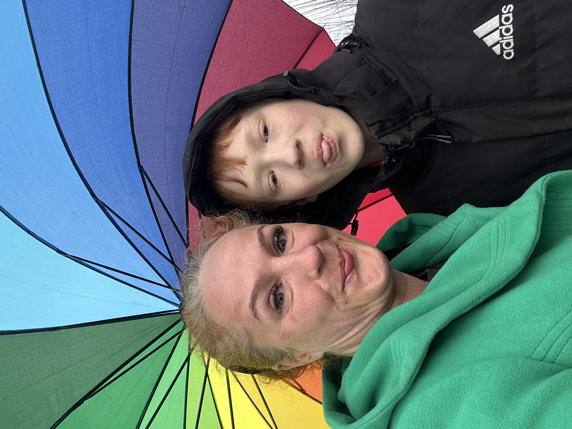 A photograph shows a woman and a child posing under a rainbow umbrella. The woman, in the foreground, has blonde hair and is wearing a green hoodie. Behind her, a child with red hair is making a face. The child is wearing a black Adidas jacket. The background is indistinct. The location is unspecified, and there is no indication of Chaikovsky, Russia.