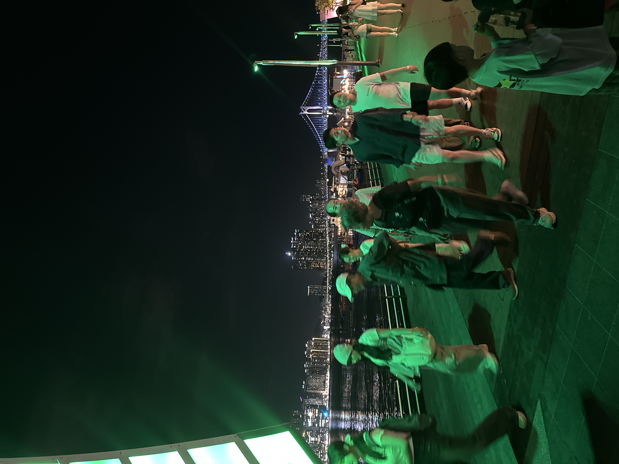 This image depicts a nighttime scene along the Busan North Port waterfront in Busan, South Korea. A group of people are walking along a promenade illuminated by green lights. In the background, the city skyline is visible with numerous high-rise buildings and the Gwangan Diamond Bridge illuminated with colorful lights. The water of the port reflects the city lights, creating a vibrant atmosphere. Several individuals are captured in motion, suggesting a leisurely stroll. The overall ambiance is one of urban nightlife and waterfront activity.