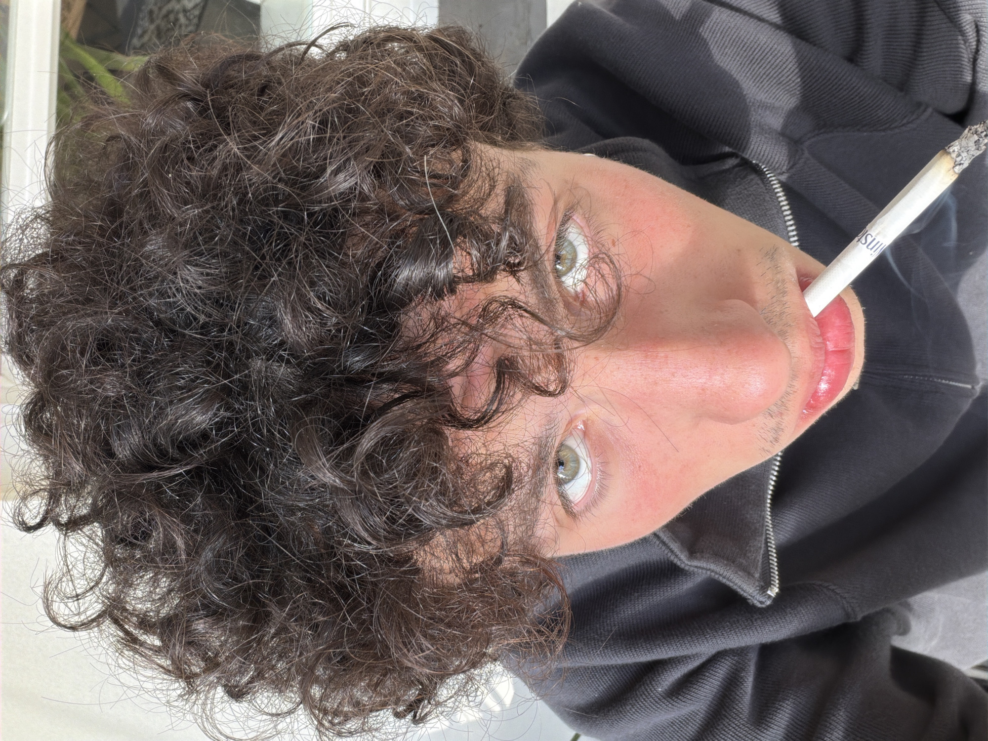 A selfie photograph shows a person with dark curly hair and light green eyes looking up at the camera, with a lit cigarette held in their mouth. The cigarette has a filter with text on it. The person is wearing a dark gray zippered hoodie. The photo was likely taken outdoors. The location in the city of Tel-Aviv, Israel is not determinable.