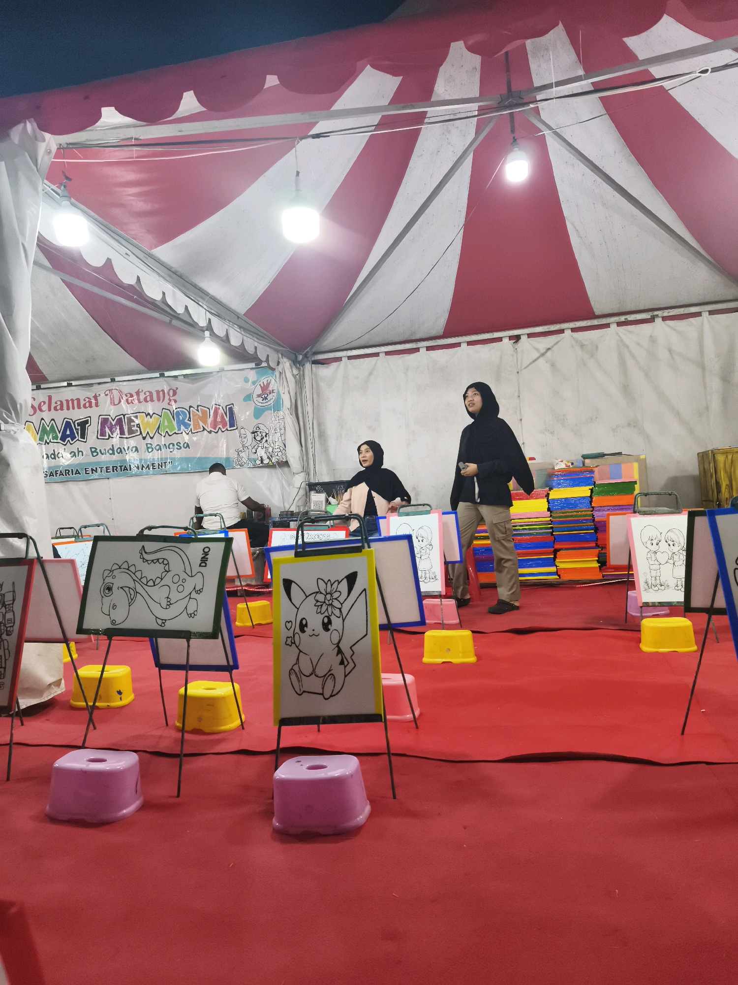 The image depicts an indoor event space, likely a tent, with a red and white striped canopy overhead. The floor is covered in a red carpet. Several easels are set up, each displaying a coloring page with a black and white outline drawing. The drawings include a dinosaur, Pikachu, and two children holding hands. There are small plastic stools in pink and yellow scattered around the easels. In the background, a banner reads "SELAMAT DATANG" (Welcome) and "MAHAT MEWARNAI" (Coloring Contest) in colorful text. Two women wearing hijabs are present, one in the background near the banner and another standing to the right, looking towards the easels. A man in a white shirt is seated behind a table in the background. To the right of the easels, stacks of colorful papers or mats are visible. The overall scene suggests an art or coloring activity event. The banner includes the text "SAFARIA ENTERTAINMENT" indicating the organizer or location. Based on available information, this event is likely held in Palembang, Indonesia, possibly at a venue like Palembang Icon or similar shopping mall or public space that hosts such family-friendly activities.