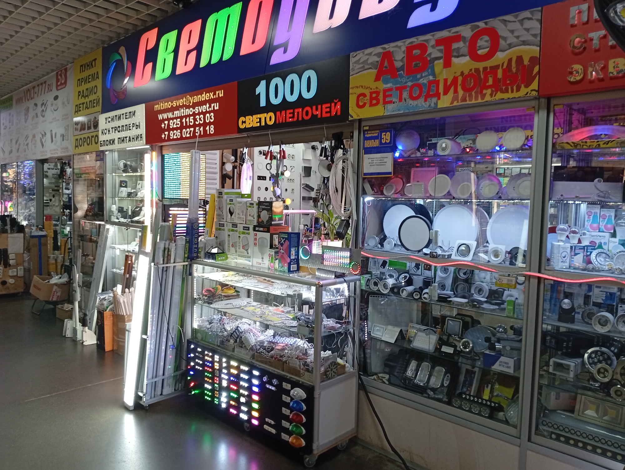 This is a medium shot of a brightly lit indoor market stall specializing in lighting and electronics. The stall is densely packed with various products, including LED lights, power supplies, and lighting accessories displayed in glass cabinets and on shelves. A prominent sign above the stall reads "СВЕТО" (meaning "light") in large, colorful, illuminated letters. Below this, smaller signs advertise services and contact information, including a website "mitino-svet.ru" and phone numbers. Another sign highlights "1000 СВЕТОМЕЛОЧЕЙ" ("1000 lighting small items"). The overall environment suggests a retail setting with a focus on illumination products.