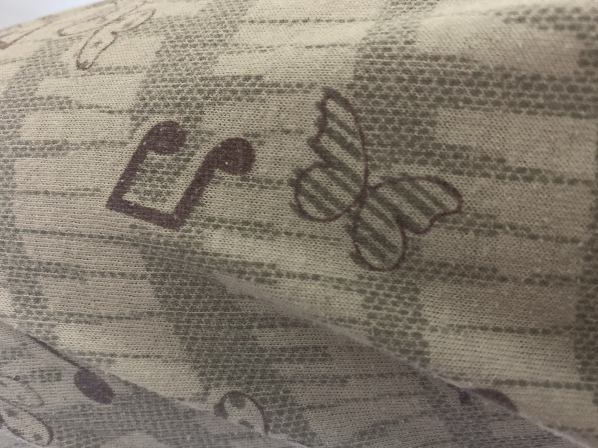 A close-up, high-angle shot captures a piece of light-colored fabric, likely a textile item such as clothing or bedding. The fabric itself is predominantly an off-white or very light beige, with a soft, slightly irregular texture consistent with a knit or woven material.
The fabric is adorned with a repeating pattern in muted tones. A subtle, underlying grid-like or checkered design in a lighter gray creates a faded background effect. Overlaid on this are distinct motifs printed in a darker brownish-purple color. These motifs include musical notes, specifically a sixteenth note (quaver) with two flags and another note with a single flag, along with stylized butterflies. The butterflies feature a striped pattern on their wings. The fabric is gently folded or draped, creating soft shadows and contours within the frame.
There are no people, activities, or events depicted. The image solely focuses on the textile's surface. No specific time of day, weather conditions, or detailed location cues are visible within the image, although the external context indicates Sofia, Bulgaria. No visible text is present; the musical notes are symbols.