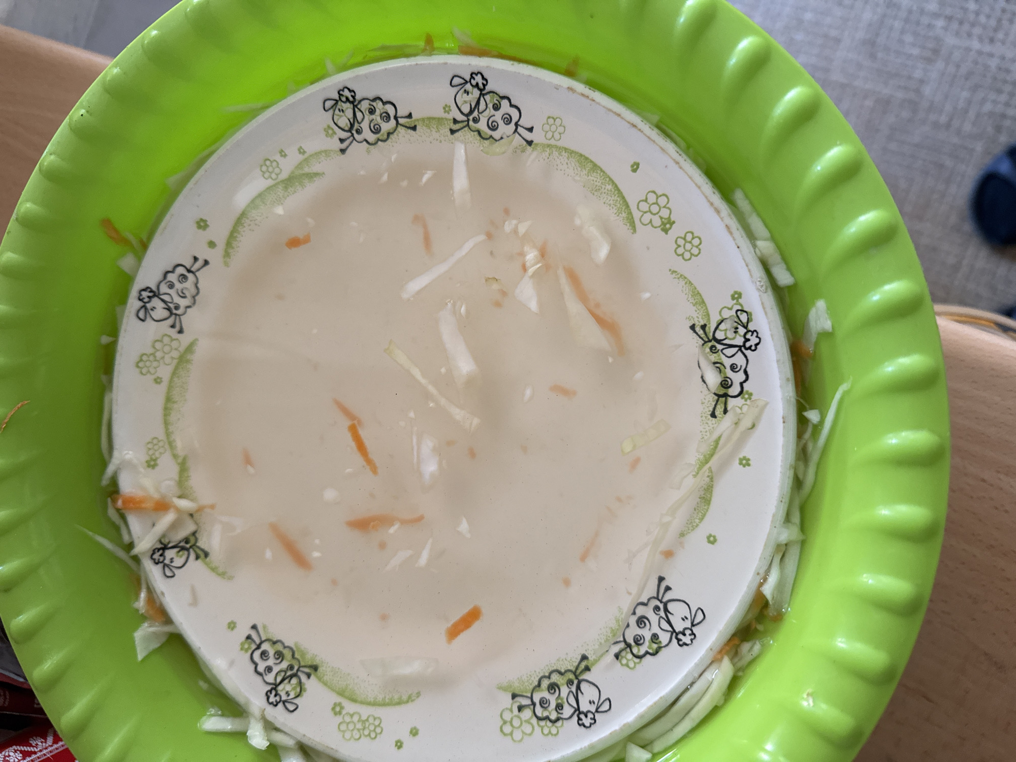 The image shows a top-down view of a white plate with a cartoon sheep design, placed inside a bright green plastic bowl. The plate contains a light-colored liquid and pieces of shredded vegetables, appearing to be cabbage and carrots. The vegetables are also visible around the rim of the plate, within the green bowl. The plate has a decorative border with small green flowers and sheep illustrations. The overall scene suggests a food preparation or presentation context. The lighting is natural, indicating it could be daytime. The background is blurred, showing a wooden surface and a hint of grey fabric, possibly a table and a chair or sofa. There is no visible text in the image.