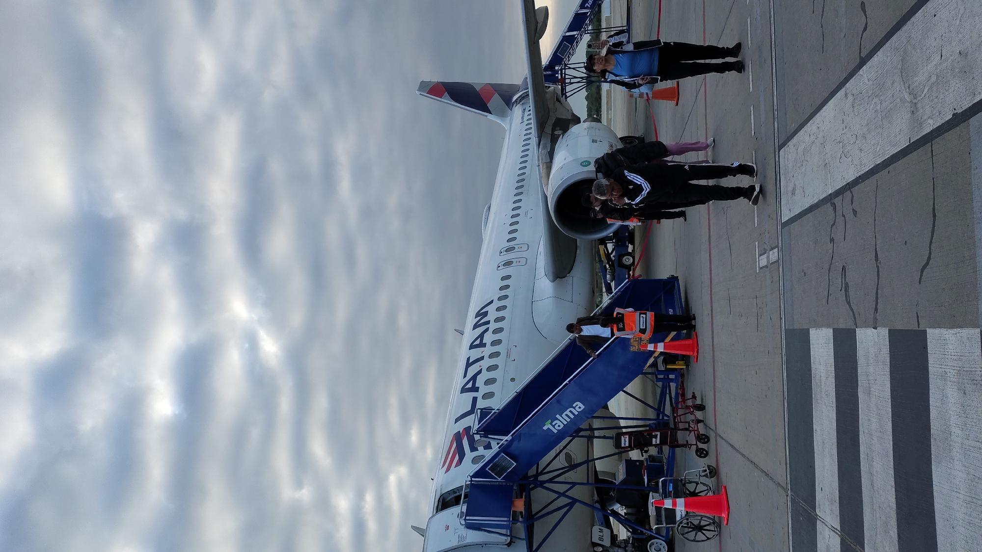 A daytime, exterior shot depicts an airplane, likely at the airport of Castilla, Peru. The aircraft, bearing the livery of LATAM, is parked on a concrete tarmac under a sky filled with stratus clouds. Passengers disembark the plane via a blue "Talma" branded boarding stair. Several individuals are present: passengers deplaning and ground personnel assisting. Orange safety cones are positioned around the boarding stairs. The location includes painted white and black stripes on the tarmac.