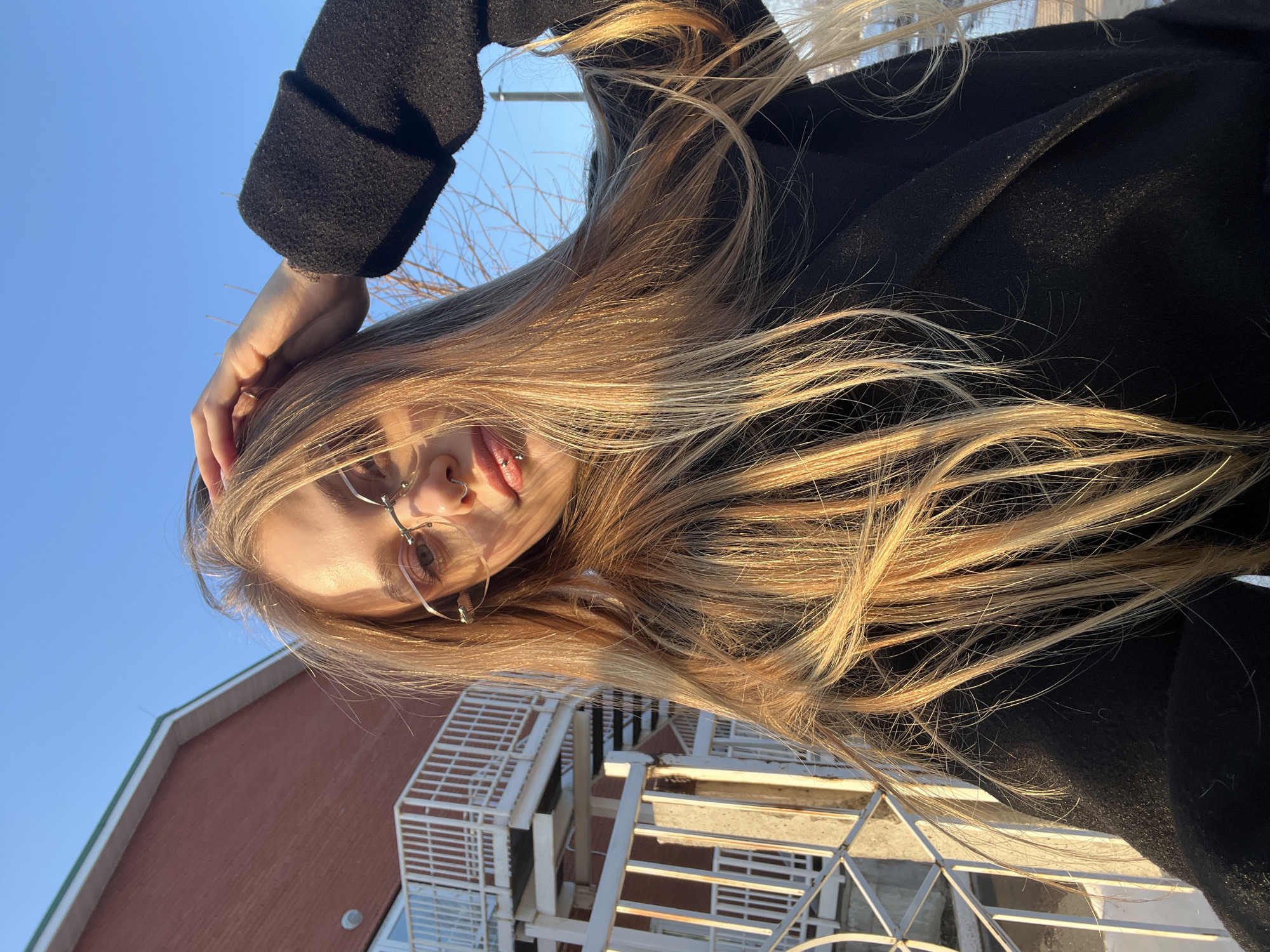 The image presents a selfie-style portrait of a young female subject outdoors in the city of Foki, Russia, under clear blue skies. The subject has long, straight, blonde hair, brightly illuminated by direct sunlight. She wears rimless eyeglasses with small, heart-shaped or wing-like metallic embellishments at the top outer corner of each lens. Her face features a labret piercing below the lower lip and a ring piercing on the left nostril. She is wearing a dark, possibly black, textured coat. Her left hand is positioned with fingers touching the hair on the crown of her head. In the background, to the left, a building constructed of reddish-brown brick with a dark green roofline is visible. Attached to this building is a prominent white metal exterior staircase and railing system. Patches of snow are present on the ground in the lower right portion of the background, and bare tree branches are visible against the sky in the middle right. A utility pole is faintly discernible in the far right background.