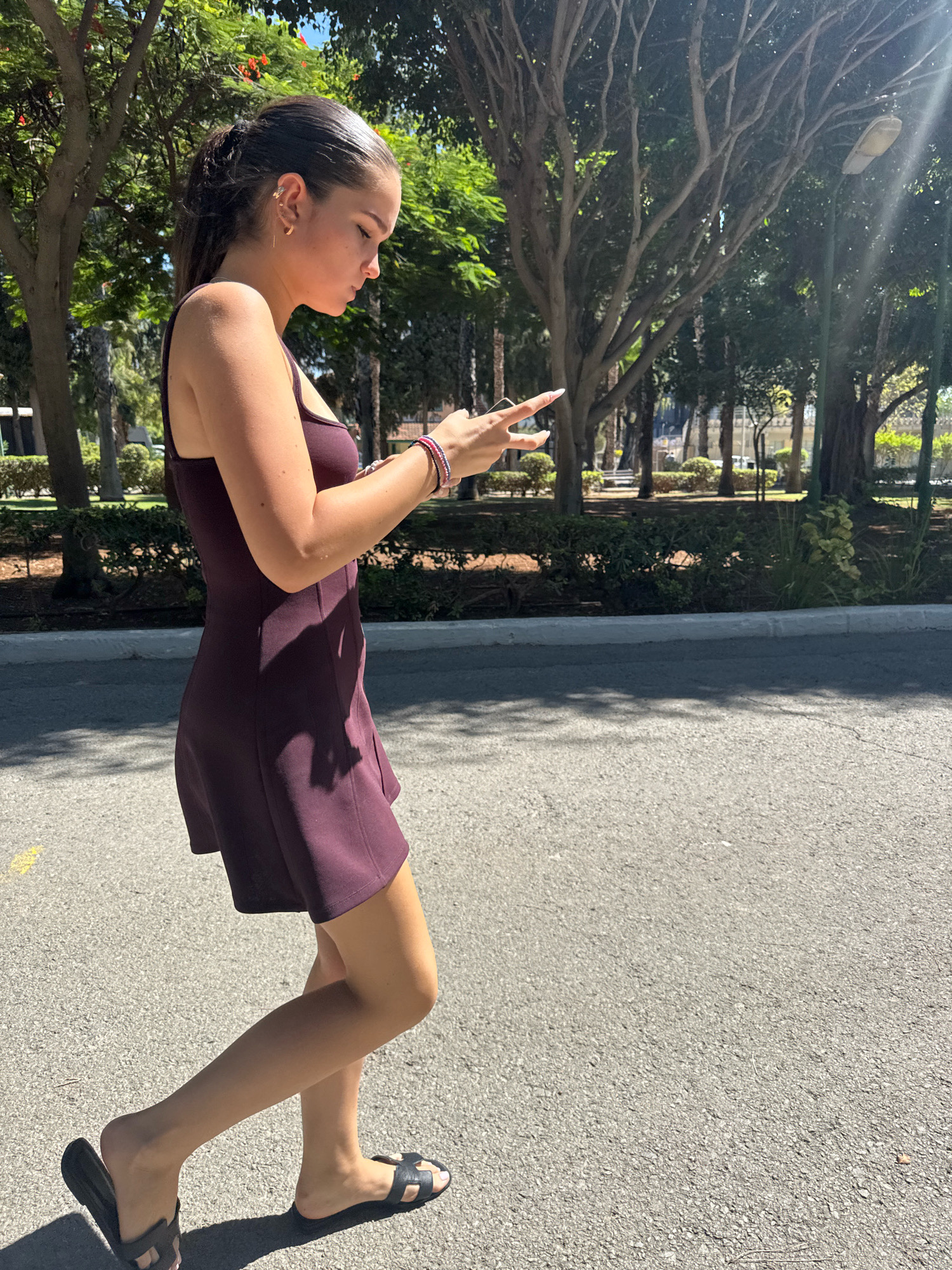A young woman with dark hair in a ponytail, wearing a dark burgundy dress and black slides, walks on an asphalt path outdoors. She is looking down at her phone, held in her hands. She is wearing bracelets on her left wrist. The background features lush green trees and bushes, suggesting a park or a tree-lined street in Limassol, Cyprus. The lighting indicates it is daytime, with sunlight filtering through the leaves.