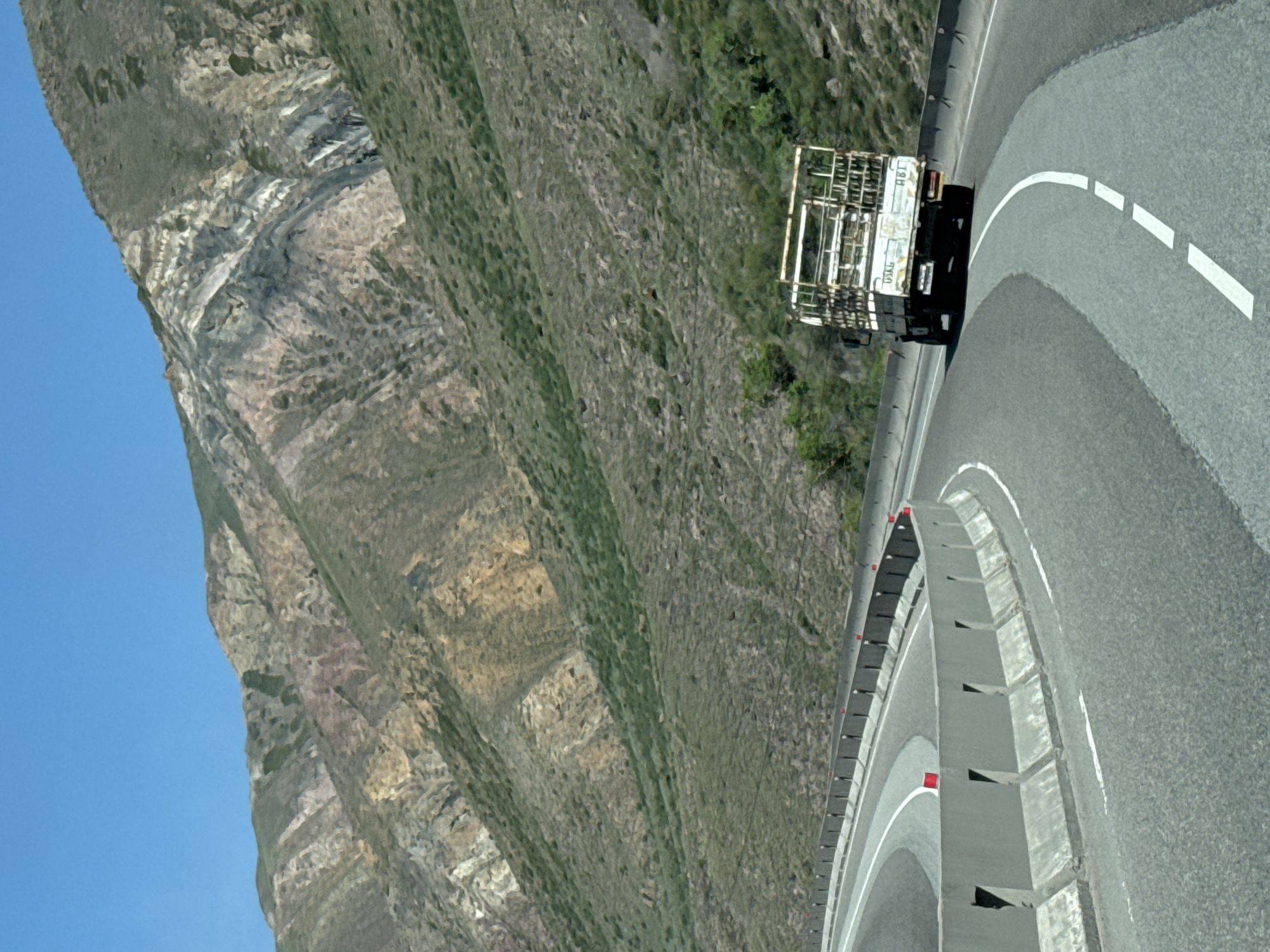 Here is a detailed description of the image:
The image shows a truck on a curved section of road. The road is marked with a solid white line and dashed white lines. A concrete barrier runs alongside the road. A steep, rocky mountain slope rises to the left of the road. The sky is blue. Based on the location provided, this scene is likely in the mountainous region near Krasnyy Most, Kyrgyzstan. The image captures a vehicle navigating a mountainous highway with safety barriers in place.