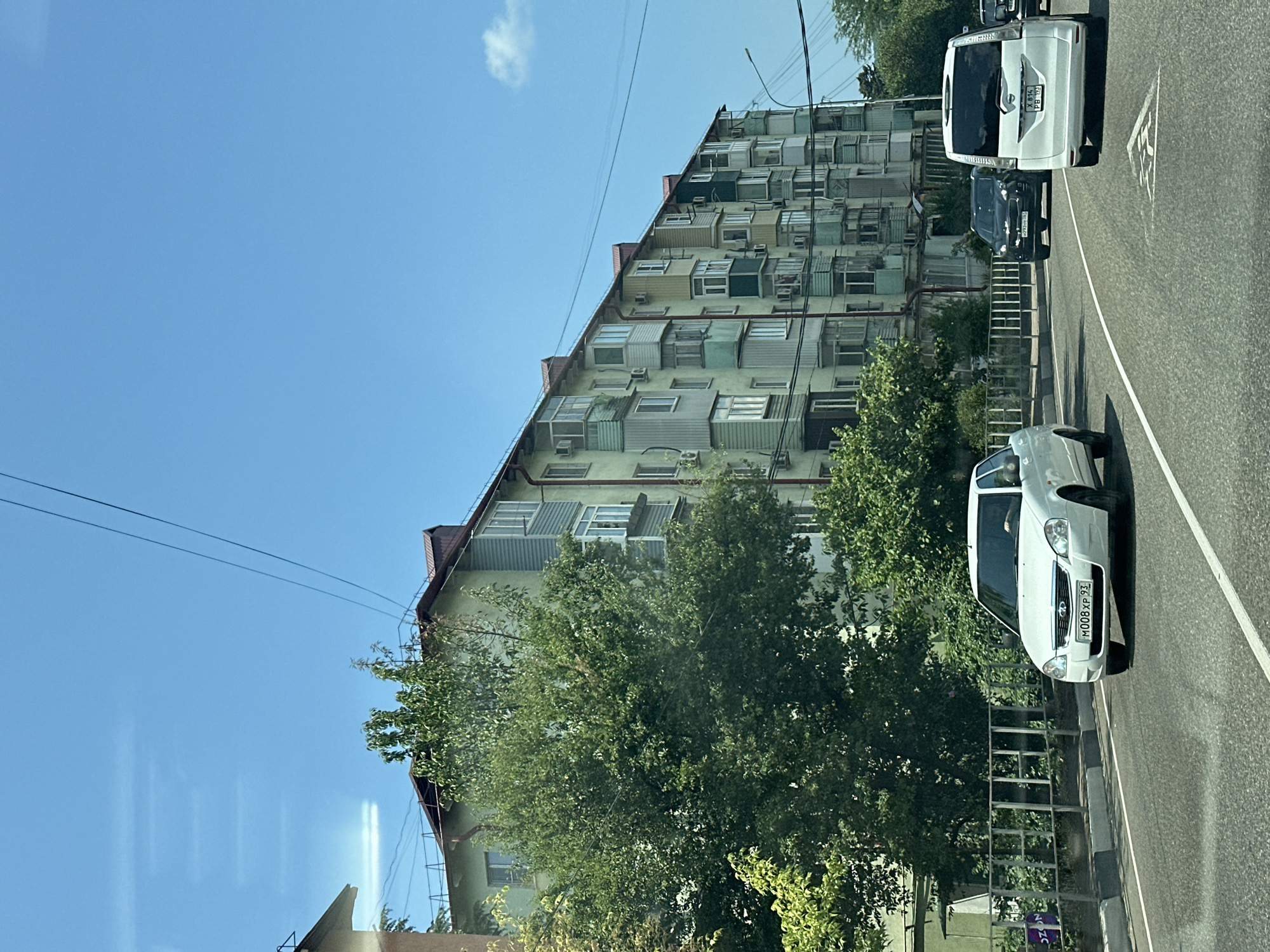 A long, multi-story apartment building with a red-tiled roof is visible on the right side of the image, painted a pale green with colorful balconies and window frames. The building appears to be in an urban setting, with a clear blue sky above and some electrical wires crossing the frame. In the foreground, a grey asphalt road with white lane markings leads into the distance. Several cars are driving on the road. A white sedan is in the left lane, with its license plate reading "M008XP 93". A black SUV is in the right lane, followed by a white minivan with the license plate "X 814 PB 193". Lush green trees and a metal fence line the side of the road, separating it from the building. The overall scene suggests a typical day in a Russian city, possibly Tuapse, Russia, given the license plate format.