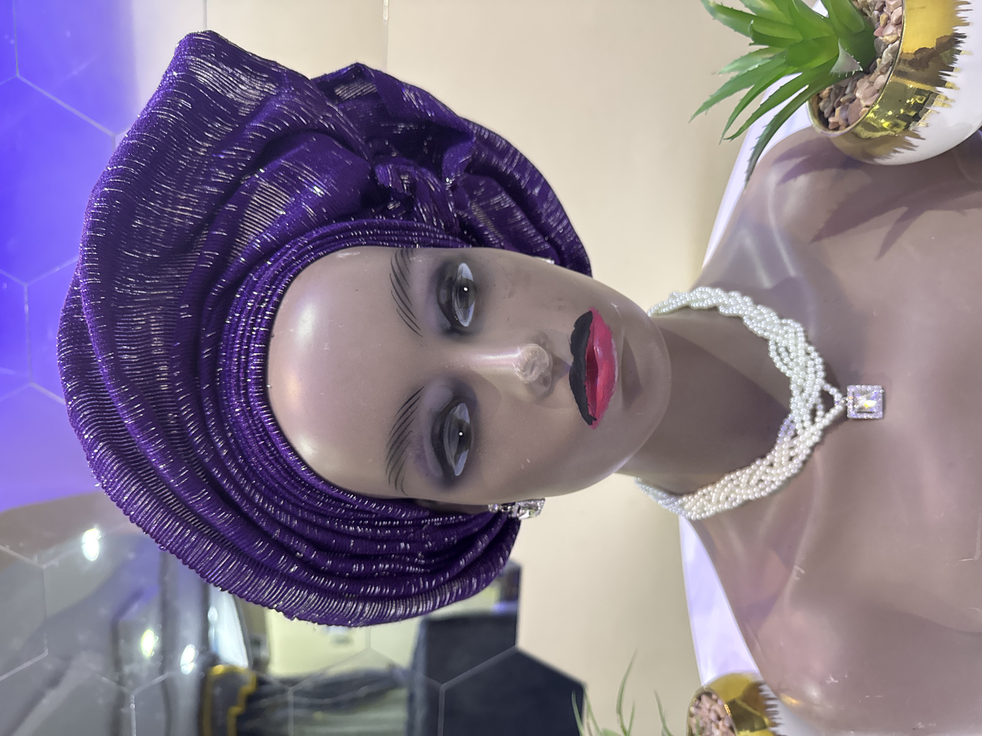 A mannequin head wearing a purple headwrap and a pearl necklace with a square pendant is displayed indoors. The mannequin has painted facial features, including eyes, eyebrows, and lips. A small potted plant with green leaves is located on the right side of the image, near the mannequin's shoulder. The background features a blue illuminated surface on the left and neutral tones elsewhere. No specific location in Ajara, Nigeria, is discernible from the image.