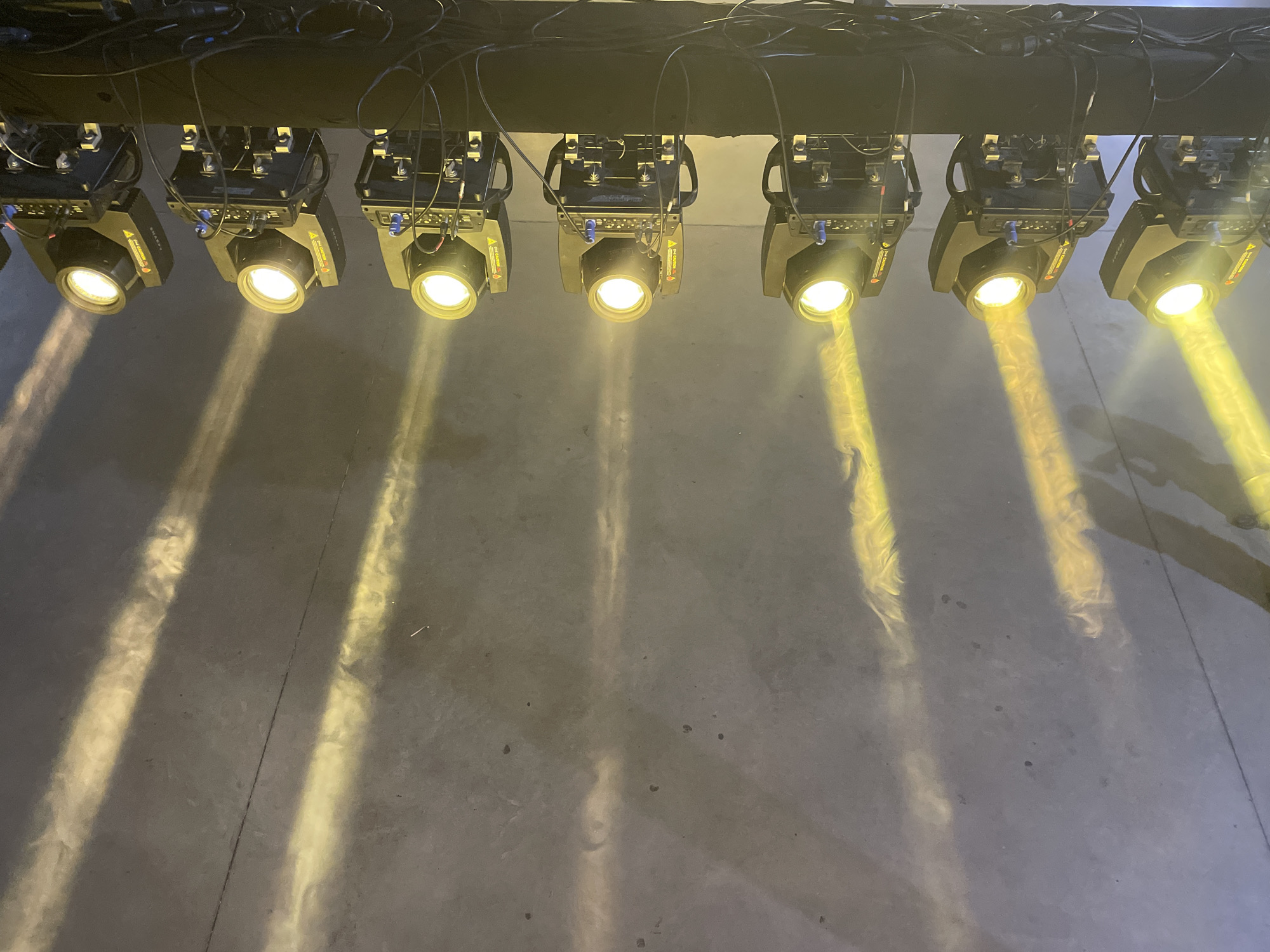 Nine bright, yellow-white spotlights are prominently featured, suspended in a horizontal line from a dark overhead truss or beam. Black electrical wires and cables are visible, connecting the lighting units and running along the support structure. Each spotlight projects a powerful beam of light downwards and slightly outwards, creating distinct, visible cones of illumination through what appears to be atmospheric haze or dust in the air.
These light beams land on a dark, concrete-like floor, forming elongated, wavy patterns of bright yellow light. The floor itself shows textural variations and a thin, dark line or seam running diagonally across the middle-right section. On the side of some of the spotlights, a red and yellow safety sticker is partially visible, bearing text such as "WARNING HOT" and "DANGER," common safety advisories for such equipment.
The scene depicts an indoor setting, strongly suggesting a stage, event venue, or studio in Buenos Aires, Argentina. The arrangement and nature of the lights indicate a professional lighting setup, possibly for an event, performance, or demonstration, with the visible light beams enhanced by an atmospheric effect. No individuals are present in the image.