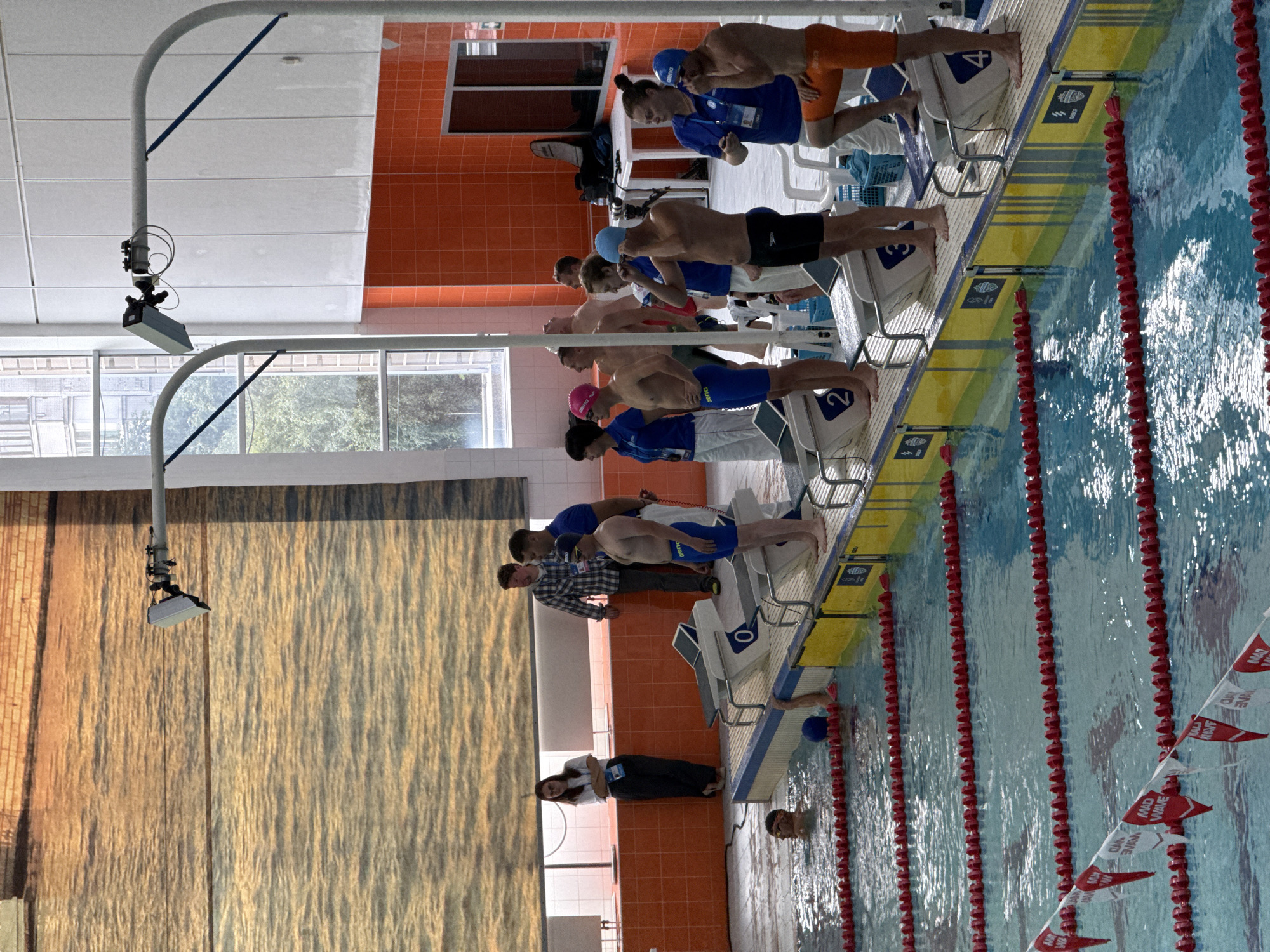The image depicts a swimming competition at an indoor pool. The pool features multiple lanes marked with red lane ropes and flags. Several swimmers are visible, some in the water, and others on the starting blocks, preparing to race. Officials and spectators are also present, observing the event. The background shows an orange-tiled wall and large windows, suggesting a modern sports facility. The location is identified as the city of Sinyavino, Russia. The image captures a moment of anticipation and readiness before a swimming race.