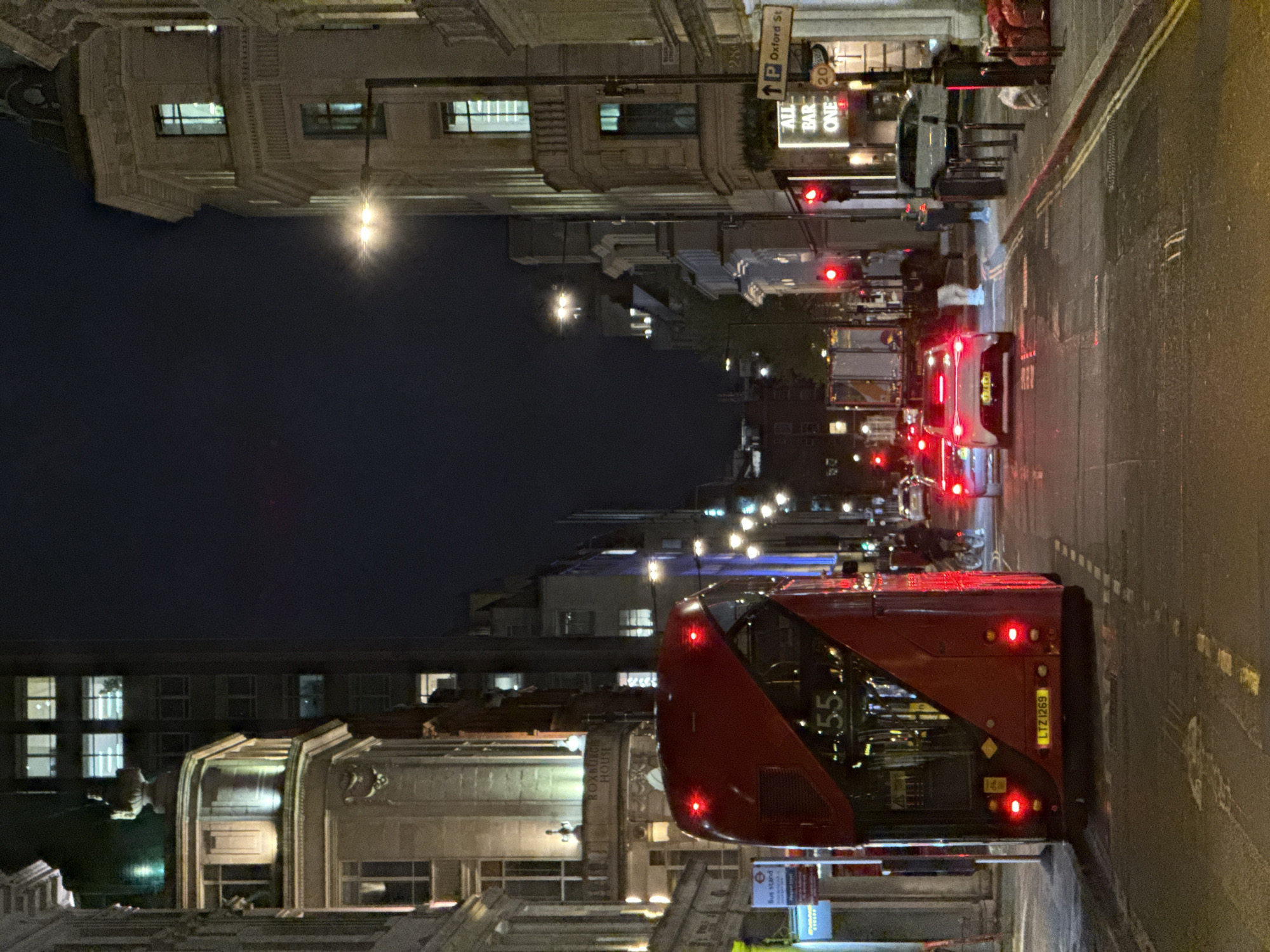 This is a night-time photograph of a bustling street in London, United Kingdom. The image depicts a red double-decker bus, a quintessential symbol of London, prominently positioned on the left side of the road. The bus's rear lights are illuminated, and its route number "155" is visible, along with its license plate "LTZ 1269".
The street appears damp or wet, reflecting the artificial lights from vehicles and streetlights, creating a vibrant glow. Several other cars are visible further down the road, with their brake lights illuminated, suggesting that traffic is halted, likely at a red light which can be seen in the distance.
Along both sides of the street, multi-story buildings with illuminated windows line the urban landscape. Notable text visible on street signs and building facades includes "Oxford St" and "ALL BAR ONE" on the right side of the road. On the left, another building has "ROXBURGHE HOUSE" inscribed, and a "Bus stand" sign is discernible near the bus. The dark sky confirms the night-time setting, with artificial light sources providing the primary illumination. The scene conveys the busy yet atmospheric character of a city street after dark.