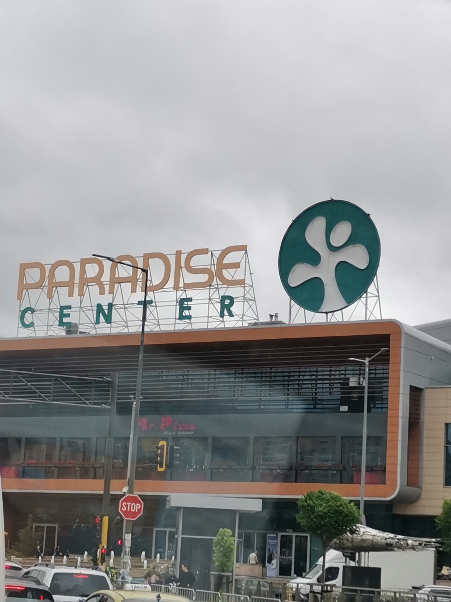 The image shows a wide shot of the Paradise Center shopping mall in Sofia, Bulgaria. The mall has a modern architectural style with large glass windows and orange trim. Above the entrance, the words "PARADISE CENTER" are prominently displayed in large, orange and green letters on a white lattice structure. To the right of the sign, a large, teal-colored circular logo with a white abstract human-like figure is visible. In the foreground, several cars are stopped at an intersection, with a red stop sign clearly visible. The sky is overcast and gray. The overall scene depicts a busy urban environment in front of a large commercial establishment.