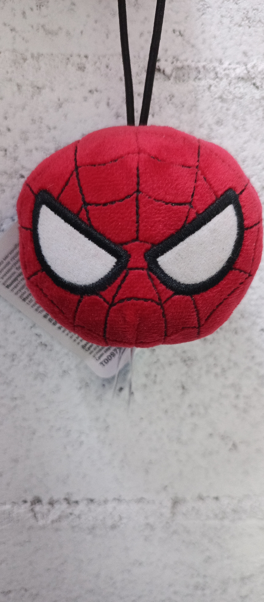 A close-up, eye-level, vertical shot shows a round, plush Spiderman mask ornament against a mottled, light-grey, textured background that appears to be a stone or concrete wall. The mask is primarily red with black web detailing and two large, white, oval-shaped eye lenses outlined in black. A black cord, split into two strands, extends from the top of the mask and disappears out of frame, suggesting it's a hanging ornament. A white tag with printed text is partially visible on the left side of the mask. The lighting is neutral, and the focus is sharp on the mask. The image is devoid of any context about its location in Bugry, Russia.