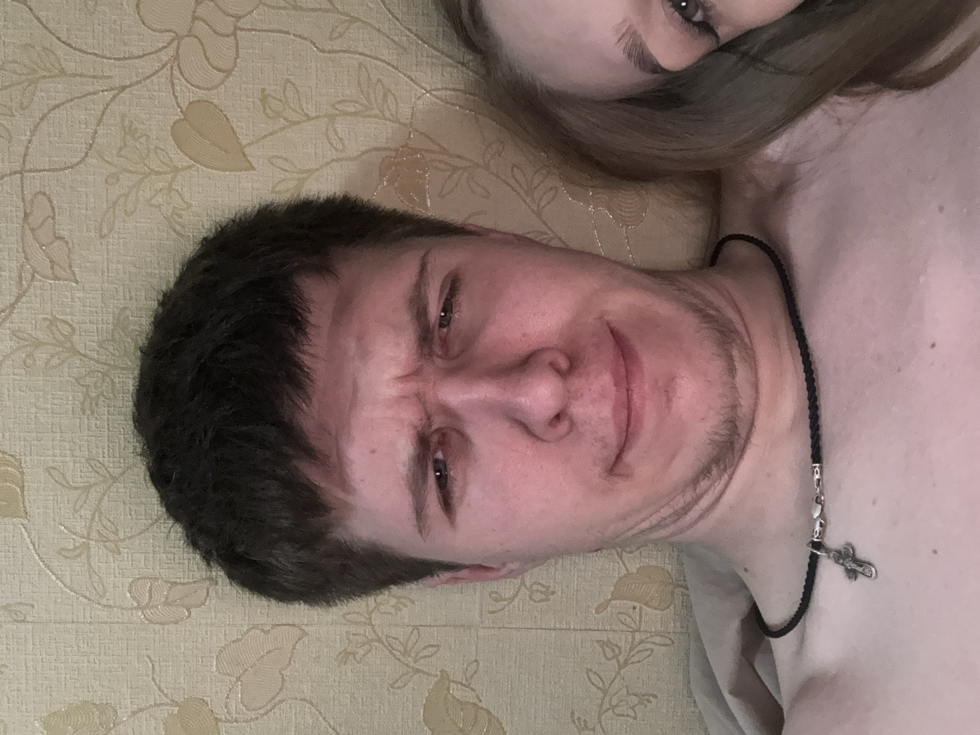This close-up image features two individuals, a man and a partially visible woman, lying down indoors in what appears to be Chaikovsky, Russia. The man, positioned prominently in the foreground, has short dark hair and light stubble. He looks directly at the camera with a furrowed brow and a slight downturned mouth, conveying a quizzical, skeptical, or perhaps slightly uncomfortable expression. He is bare-chested and wears a braided black cord necklace with a silver cross pendant.
The woman is only partially visible in the upper right of the frame, showing one eye, an eyebrow, and strands of her light-colored hair. Her expression is not discernible. The background consists of light-colored wallpaper featuring an embossed floral and leafy pattern, some elements of which have a subtle shimmer. The lighting suggests an indoor setting, possibly illuminated by artificial light. There is no visible text in the image.
