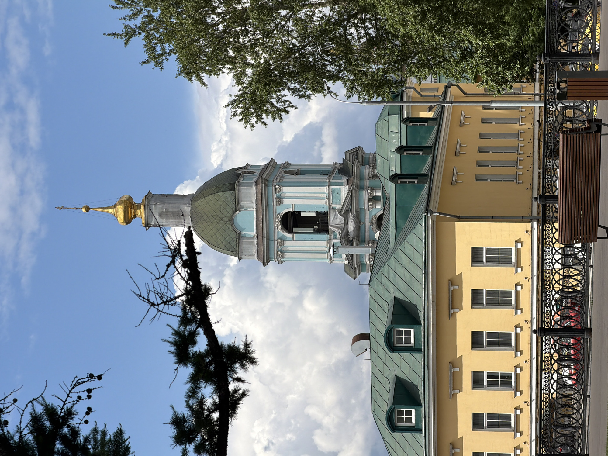 The image captures a daytime view of a church in Podolsk, Russia. The church tower, with its green dome and gold spire, stands prominently against a backdrop of a partly cloudy blue sky. A yellow building with a green roof is adjacent to the church. Trees with green foliage frame the upper portion of the scene. A section of a black metal fence and a park bench are visible in the foreground.