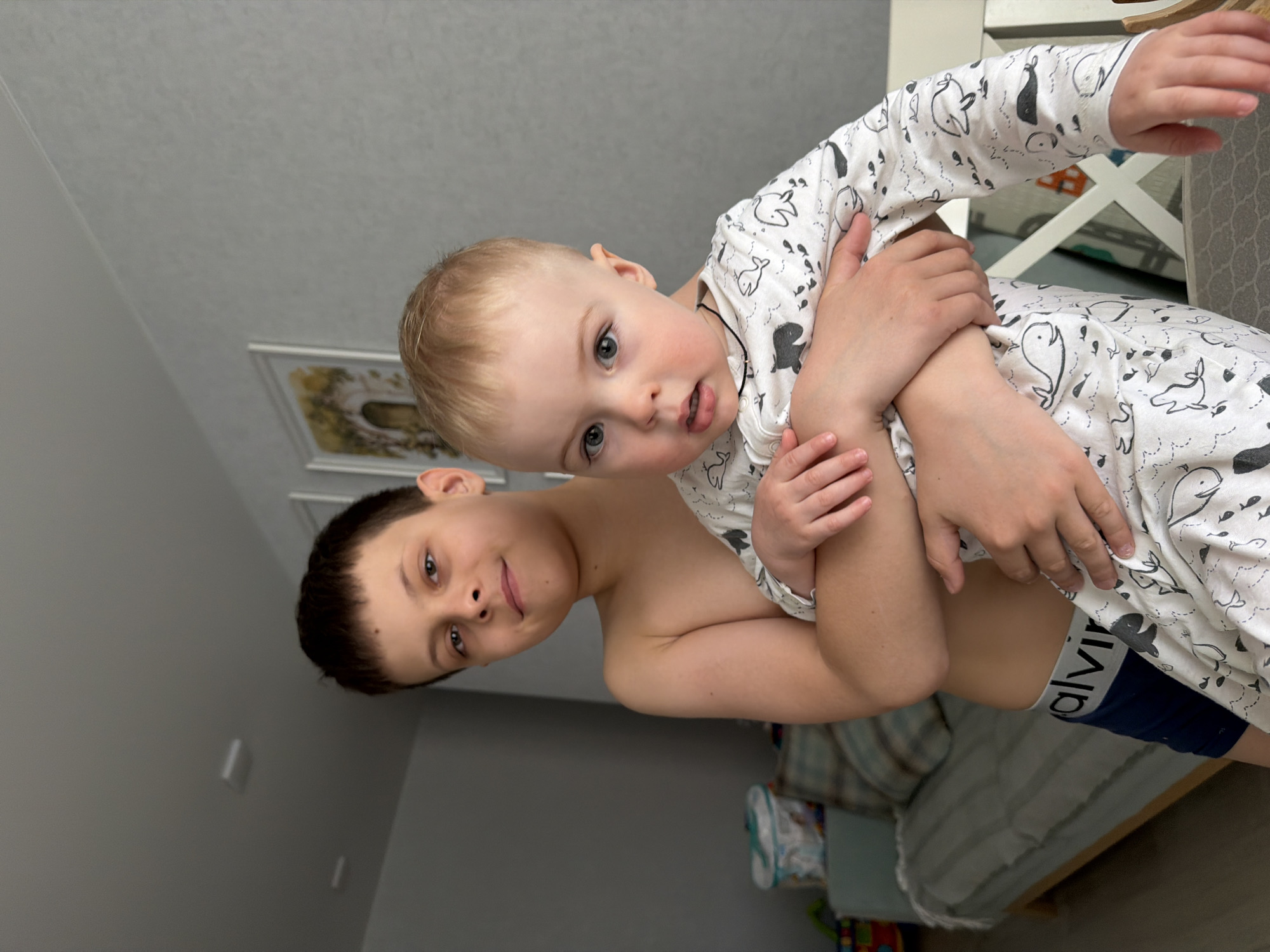 Two boys are visible indoors. An older boy, shirtless, is holding a younger boy wearing a white and black patterned pajama set. The younger boy is also wearing dark blue underwear with "Calvin" visible on the waistband. The background includes a gray wall, framed pictures, and furniture. There's no information to verify that the image was taken in the city of Votkinsk, Russia.