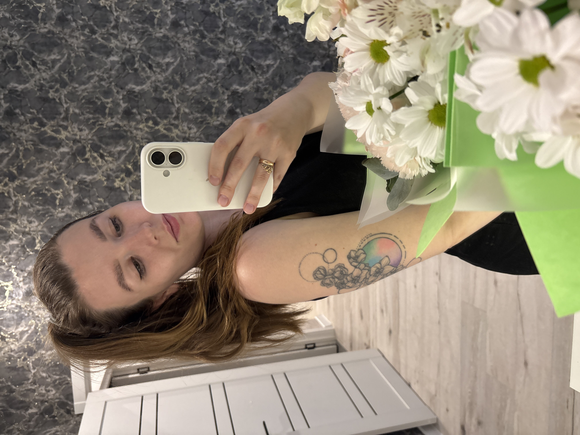 A woman is taking a selfie with a bouquet of white flowers. She is holding a white smartphone in her right hand. She has a tattoo on her left arm featuring flowers and a rainbow-colored circle. She is wearing a gold ring on her finger. The background shows a grey patterned floor and a white cabinet. The location is unknown, but the prompt suggests it might be Izhevsk, Russia.