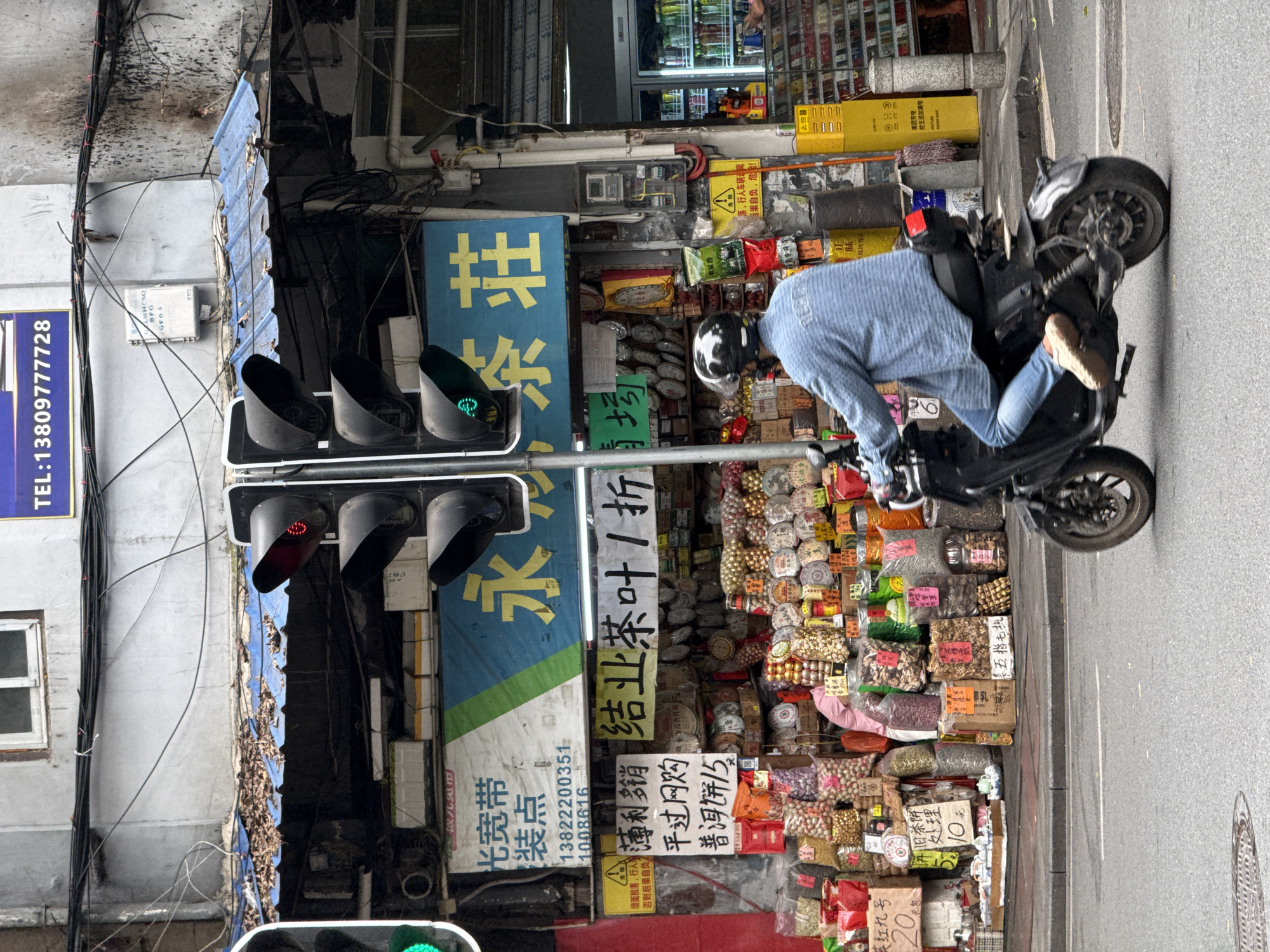 The image captures a bustling street scene in Liwan District, China. A person on a black scooter or motorcycle is the focal point, positioned on the right side of the frame, seemingly riding past a storefront.
The storefront is cluttered, showcasing an assortment of goods spilling onto the sidewalk. Packaged snacks and tea products are densely arranged. Signage is visible in Chinese characters, including business names and promotional offers. A traffic light stands on the left, with a red light illuminated at one level.
The urban environment is characterized by utility poles and wiring.