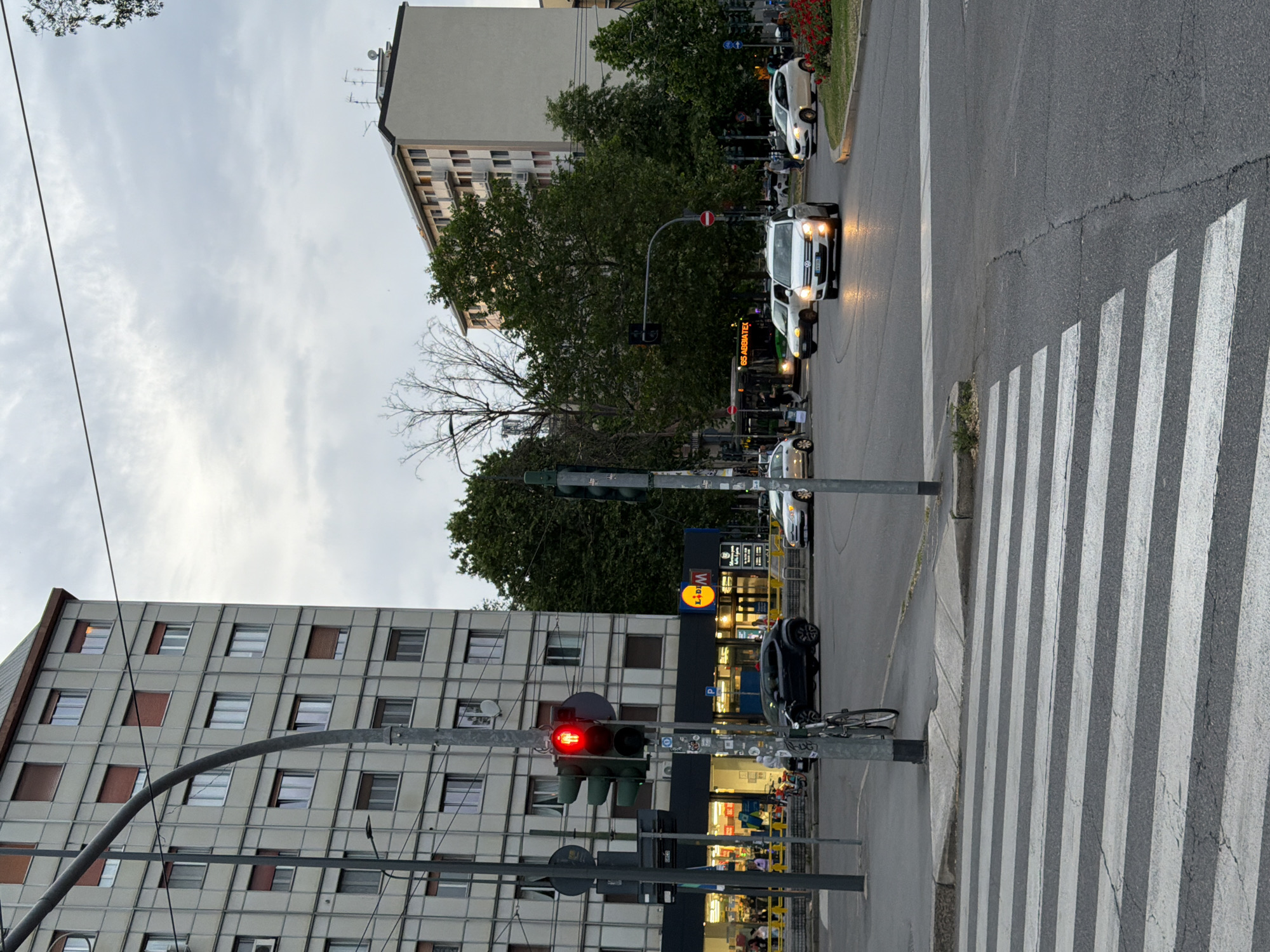 The image depicts a street scene in Milan, Italy, at an intersection with a crosswalk. The traffic light is red. On the right side of the road, vehicles including cars and a bus are stopped. A LIDL supermarket is situated on the corner. Buildings are located in the background. Pedestrians are visible.