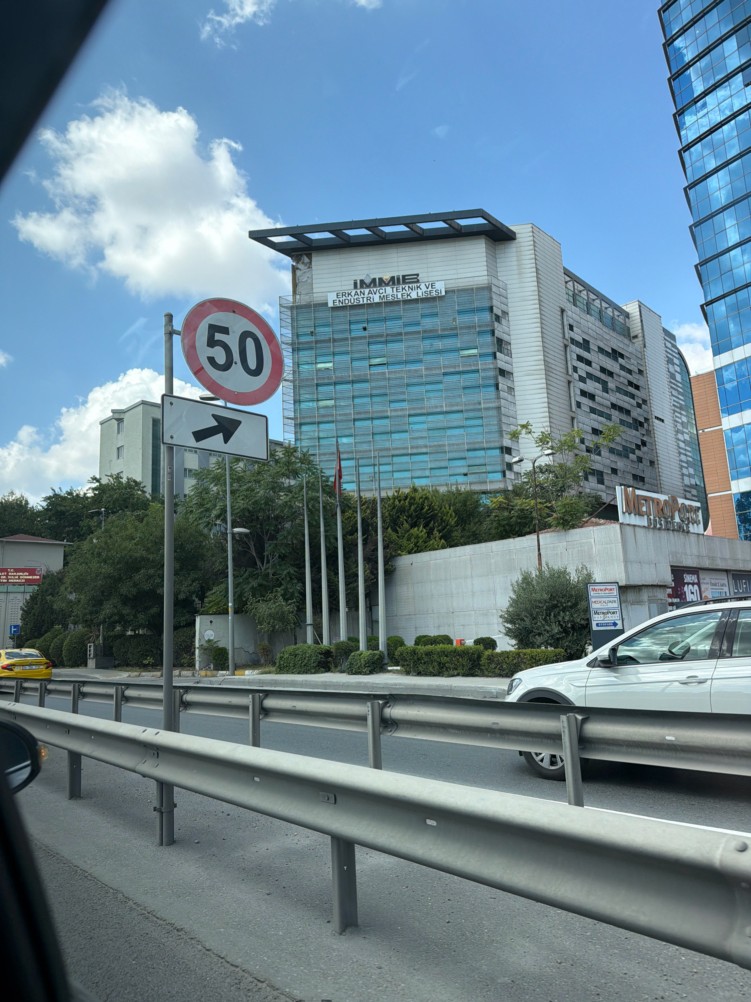 The image captures a street scene in Bakırköy, Turkey, under a bright blue sky with scattered clouds. In the foreground, a silver metal guardrail lines a road. Beyond the guardrail, a white car is visible, its front right wheel on the road. To the left of the road, a yellow taxi is parked. A speed limit sign indicates "50" and an arrow points to the right. Behind the road and signs, a multi-story building complex is prominent. The larger building on the right has a facade of reflective blue glass, while the building on the left is a lighter color with glass windows and a dark-colored structure on its roof. Text on the lighter building reads "IMMIE ERKAN AVCI TEKNIK VE ENDÜSTRI MESLEK LISESI". Trees and shrubs line the area between the road and the buildings. Another sign for "METROPORT BUSINESS" is visible on a lower section of the building.