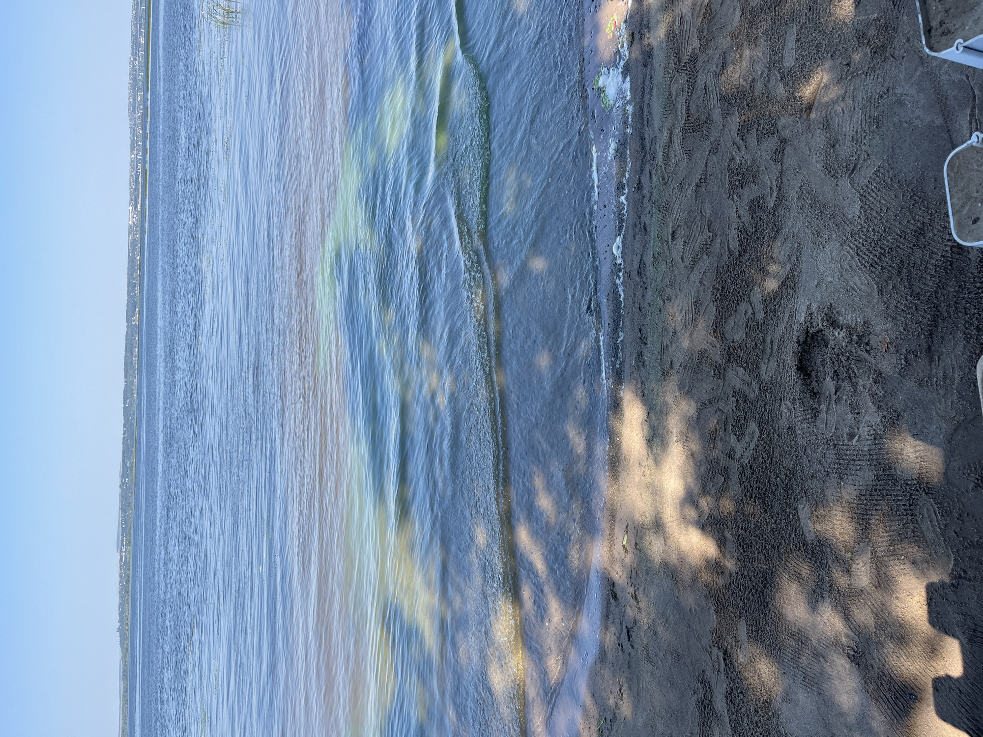This is a close-up, eye-level shot of a body of water, likely a lake or river, bordering a sandy shoreline. The water exhibits a distinct color gradient, with a reddish-brown hue near the shore transitioning to a clearer, blueish-green in the distance. Ripples and waves are visible on the water's surface, and shadows of foliage are cast upon the wet sand of the shoreline. The background shows a distant landmass with vegetation and structures, under a clear blue sky. The scene appears to be located on the outskirts of Shumeika, Russia.