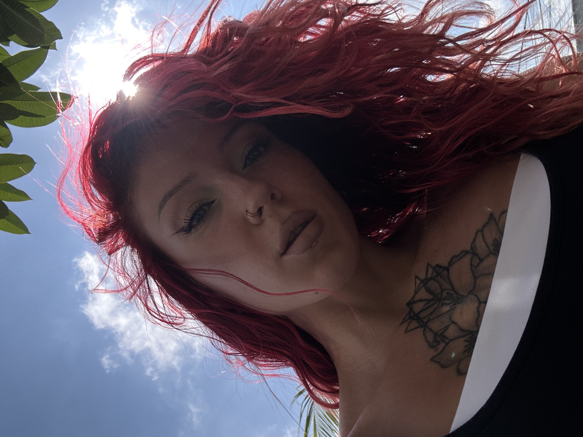 A low-angle, outdoor, selfie-style shot captures a person with vibrant red, wavy hair against a bright blue sky. The sun is shining directly, creating a lens flare that obscures part of the sky and highlights the person's hair. The person's face is turned towards the camera, with their gaze directed slightly off-center. They are wearing makeup, including eyeliner and mascara, and have a nose ring and a lip ring. A tattoo of a floral design with geometric elements is visible on their chest, just above the neckline of a black tank top. Lush green leaves are visible at the top of the frame, suggesting an outdoor, possibly tropical environment. The bottom part of the image shows the person's shoulders and the white band of their inner garment. The scene evokes a sense of casualness and self-expression, possibly taken on a sunny day in the city of North Kuta, Indonesia.