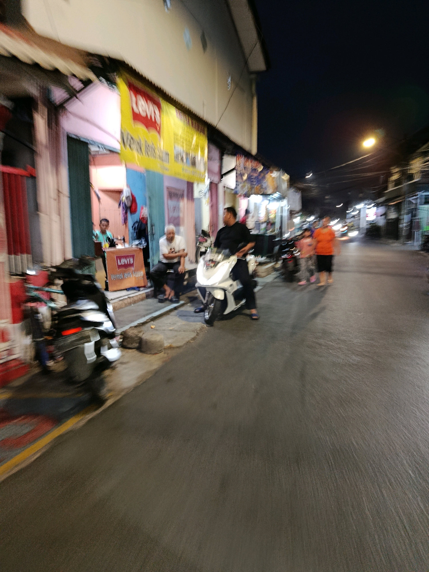 The image depicts a street scene at dusk or night in a commercial area, likely in Bekasi, Indonesia, given the text visible on signs. The foreground features two motorcycles, one black and one white, parked on the side of the road. A man in a black shirt is seated on the white motorcycle. To the left, a man in a light blue shirt stands near a shop entrance, and another man in a white shirt is seated on a dark object. In front of the shop, there is a display banner with the "Levi's" logo and Indonesian text, indicating a Levi's store or a seller of Levi's products. Further down the street, several people are walking, including a woman in an orange shirt and a child in a pink outfit, illuminated by streetlights and shop lights. The buildings lining the street appear to be shops with open fronts. The overall impression is of a bustling urban environment at night.