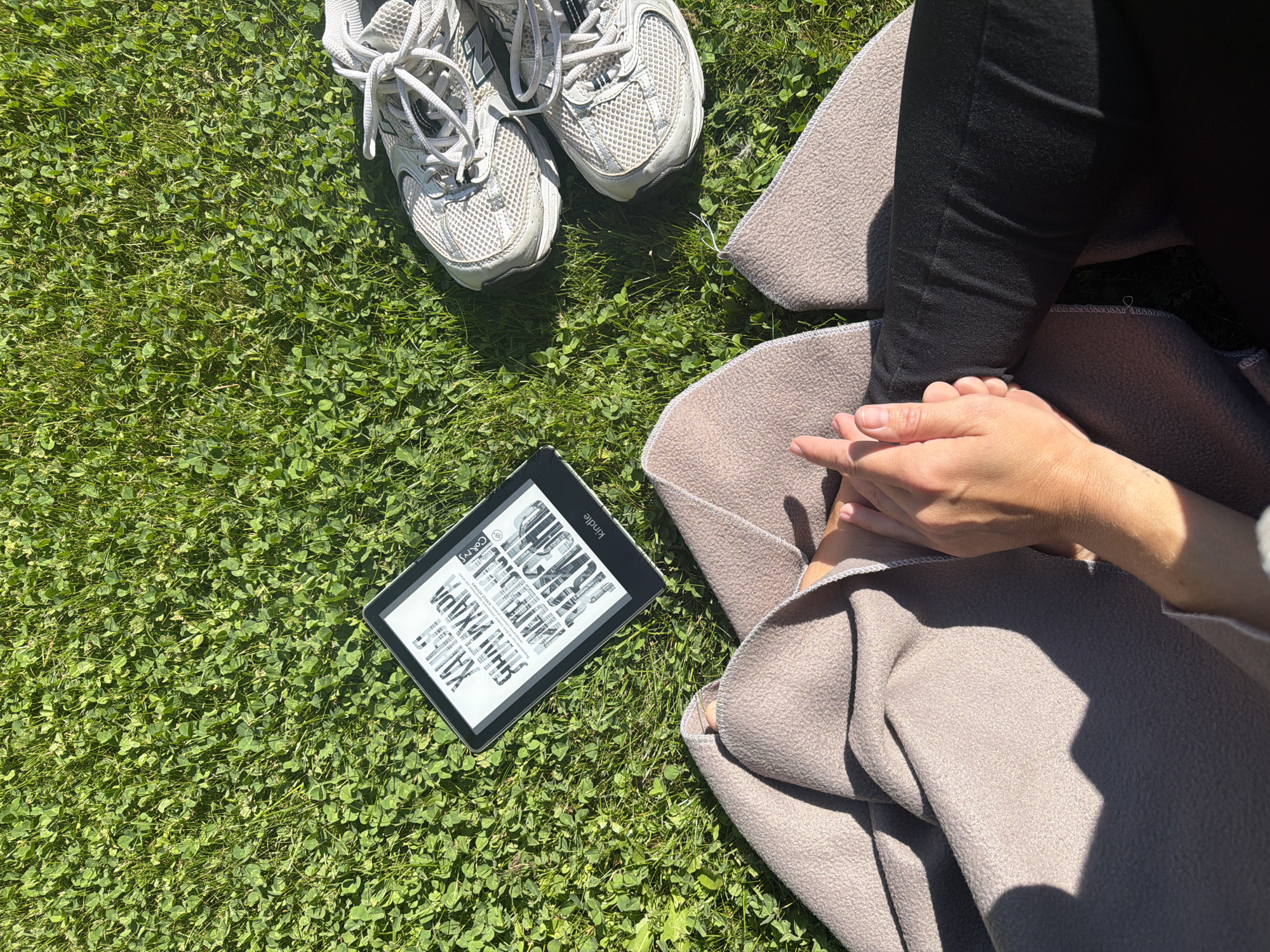 A top-down, medium shot captures a person's hands clasped in their lap, covered by a beige blanket, on a grassy area. To the right of the hands, a pair of white and grey New Balance sneakers rest on the grass. To the left of the hands and blanket is an open Kindle e-reader, displaying text on its screen. The environment is a sunny day with green grass and clover. The image composition suggests a peaceful, outdoor setting, possibly a park or backyard, in Mishutkino, Russia.