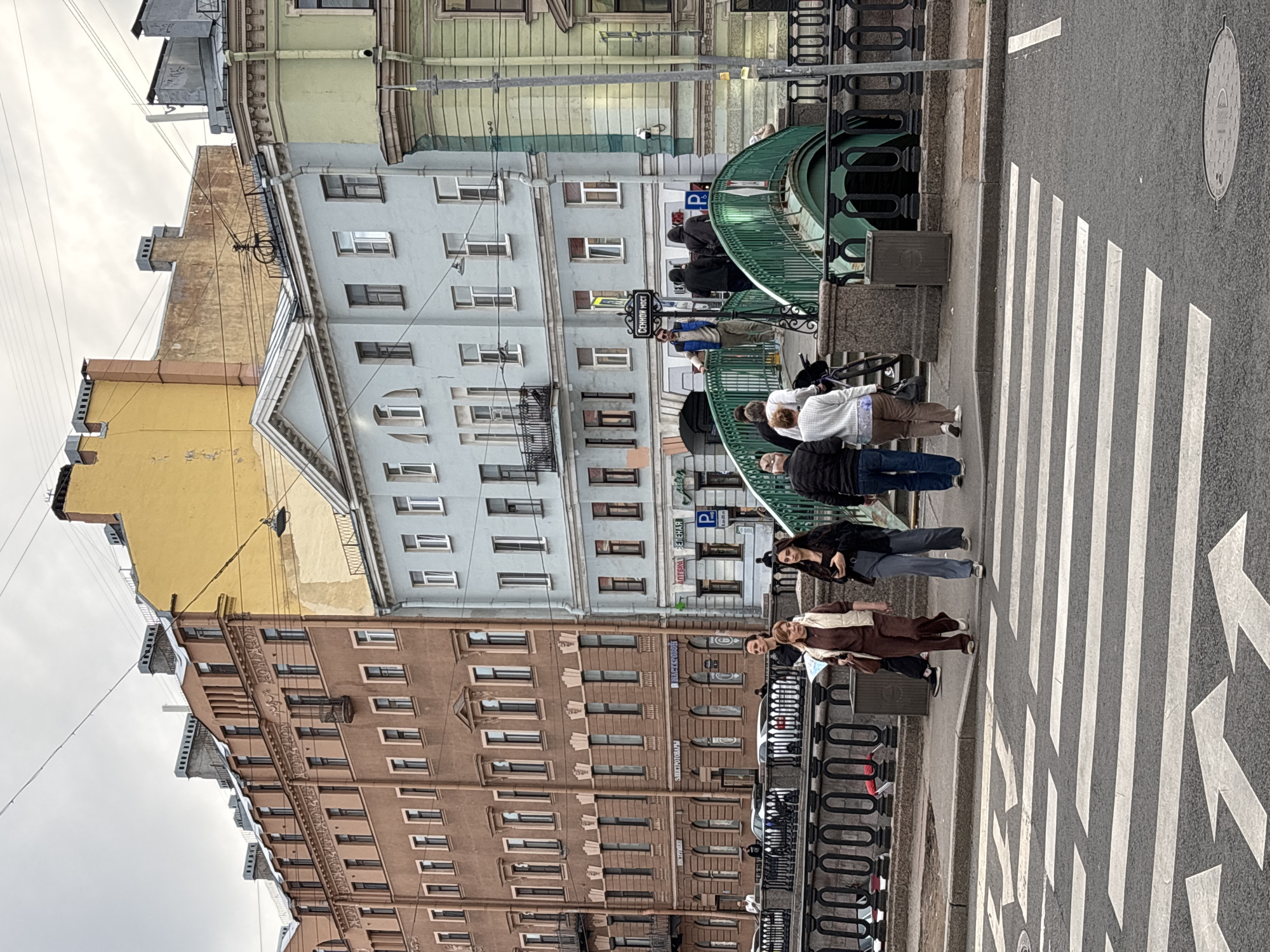 This image captures a lively street scene in Saint Petersburg, Russia, featuring historic architecture, a canal, and pedestrians. The setting appears to be a bridge or embankment area, confirmed by the text on a street sign.
**Subjects:**
Several multi-story buildings line the street. On the left, a light brown building with decorative elements stands prominently. Adjacent to it, a light blue-grey building with a distinctive pediment-like feature is visible. Further back, a pale green building can be seen. In the foreground, a wide asphalt road with a pedestrian zebra crossing occupies the right side of the frame. A dark metal railing separates the sidewalk from what appears to be a canal or river, and a curved green structure, possibly part of a bridge or an underpass entrance, is visible.
**People:**
Multiple individuals are present, engaged in daily activities. In the foreground, a person in a brown outfit and scarf walks across the crosswalk, while another in a dark jacket and jeans follows. Further along the sidewalk and near the green structure, other pedestrians are walking or standing, including a person in a white top and brown pants, and several others in various casual attire.
**Activity or Event:**
The scene depicts everyday urban life, with people walking along sidewalks, crossing the street, and moving around the bridge area. There is no specific event taking place, but rather routine pedestrian traffic.
**Notable Details:**
The time of day appears to be daytime, under an overcast sky, suggesting cloudy weather. Natural light illuminates the scene without strong shadows.
**Location Cues:** A prominent black street sign clearly displays "**СЕННОЙ МОСТ**" (Sennoy Most), identifying the location as Sennoy Bridge in Saint Petersburg. Another blue sign with a 'P' symbol and Cyrillic text, possibly "**ПЛАТНАЯ**" (Paid), is visible near the bridge, indicating a parking area. Overhead wires crisscross the sky, a common feature in older city districts. A manhole cover is visible on the street surface.
