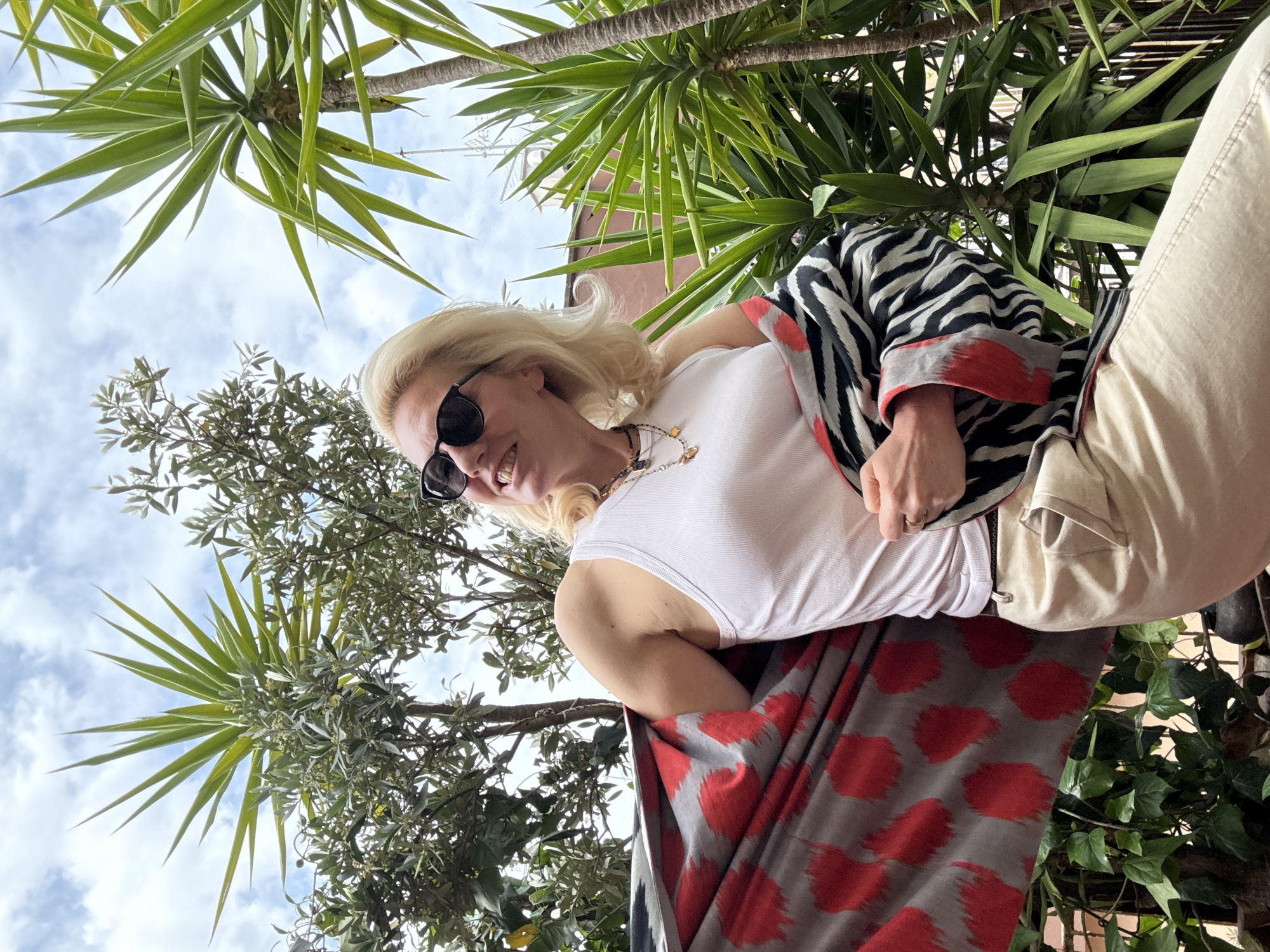 A medium, low-angle shot shows a blonde woman wearing sunglasses and a white tank top. She is also wearing a scarf with zebra stripes and red accents, and a red and gray patterned garment is draped around her. The background includes palm trees, a cloudy sky, and other green trees. Based on the foliage and architectural features, the location is likely Rome, Italy.