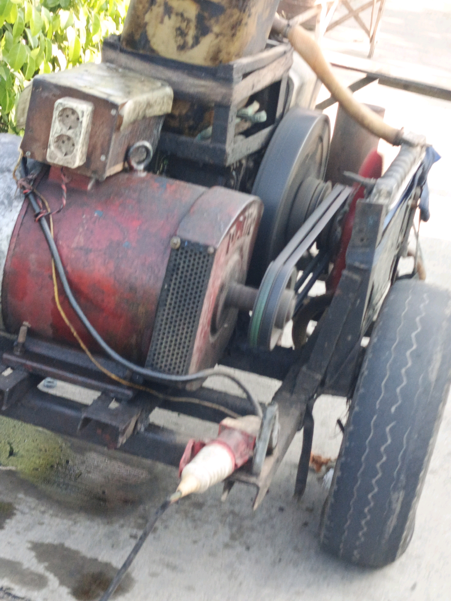 The image shows a red and yellow generator on a metal frame with a tire. It is located outdoors, on a paved surface, with some greenery in the background. The generator is connected to a power outlet and has a belt-driven system. There are no people visible in the image. The scene appears to be in a somewhat cluttered or industrial area.