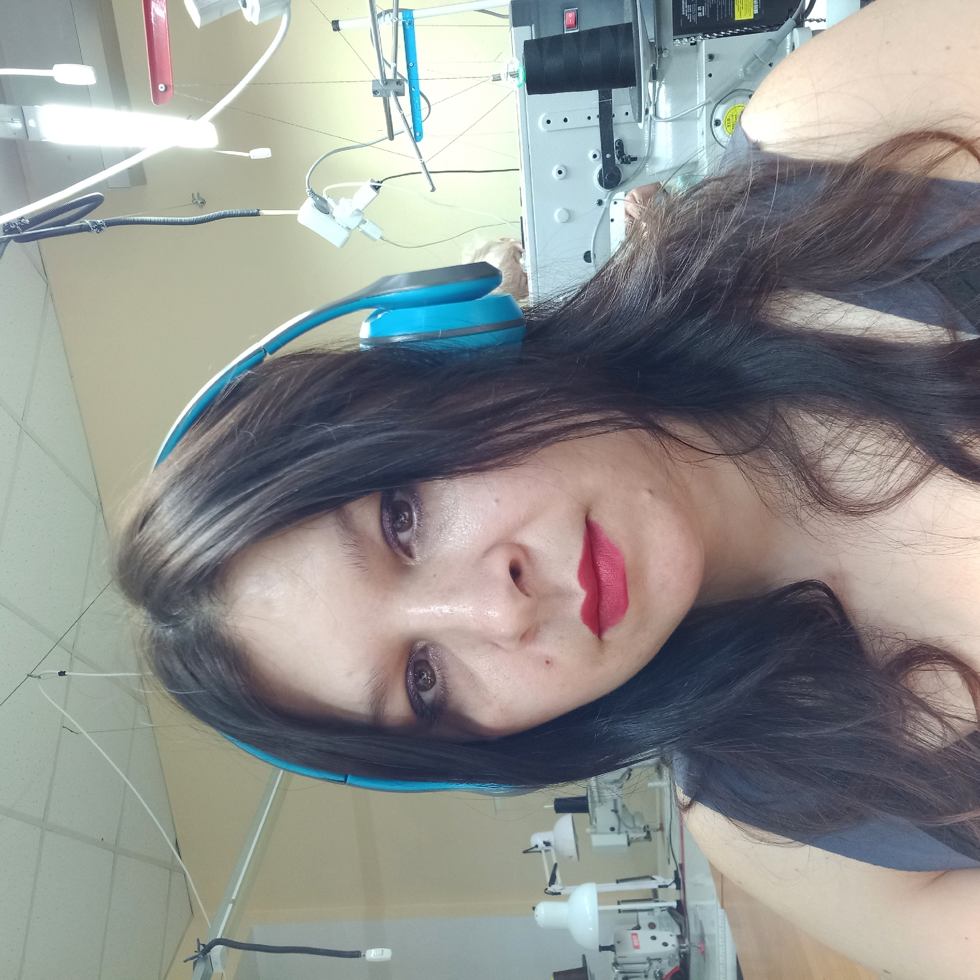 A woman with dark hair and headphones is pictured from the chest up. She is looking directly at the camera. She is wearing bright red lipstick and eyeshadow. She is in a room with sewing machines and industrial lighting. The background appears to be a workshop or factory setting. There is no indication of the location in Kropyvnytskyi, Ukraine.