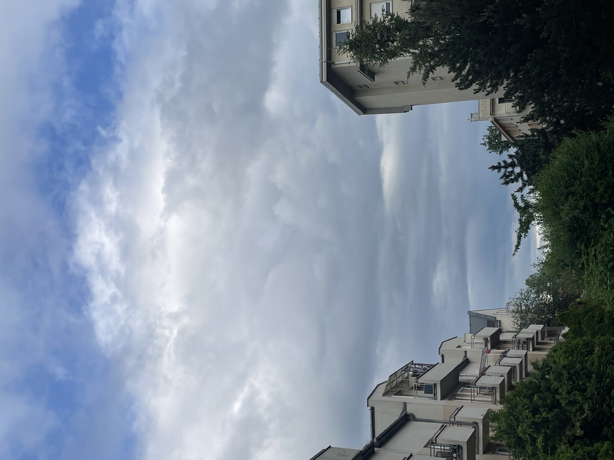 The image captures an upward view of a cloudy sky framed by modern residential buildings and trees. The sky dominates the central and left portions of the frame, displaying a mixture of light blue patches and extensive white and grey clouds, indicative of an overcast day.
Sections of two multi-story buildings are visible on the right side of the image. The upper right corner shows the corner of a light-colored building with several windows and a dark roofline, partially hidden by dark green trees. Towards the lower right, another part of a similar building features multiple balconies with metal railings, again nestled amongst lush green foliage. The architecture appears contemporary, with neutral-colored facades.
The scene is static, with no discernible human activity. The time of day is daytime, characterized by diffused light due to the heavy cloud cover. There are no specific location cues or visible text to identify the exact setting beyond a general urban or suburban residential environment. The overall mood is calm and somewhat muted due to the cloudy weather.
