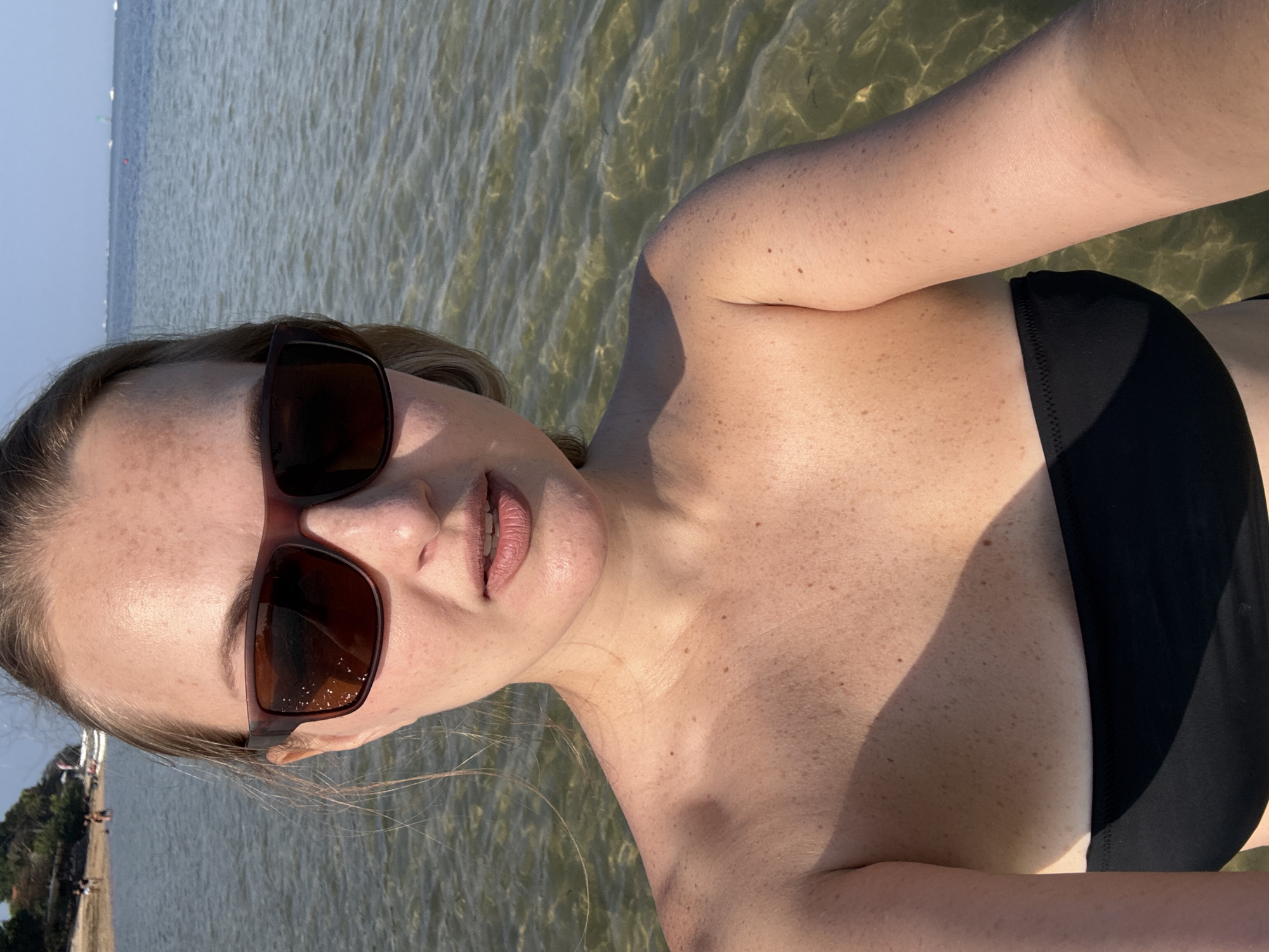A woman wearing sunglasses and a black bikini top stands in shallow, clear water. She is looking directly at the camera. Her face is illuminated by sunlight, and she has freckles across her nose and cheeks. Behind her, the water extends to the horizon, where several small boats are visible. A sandy beach with a few people can be seen in the distance on the left side of the image. The sky is clear and blue. The location is the coast of Denpasar, Indonesia.