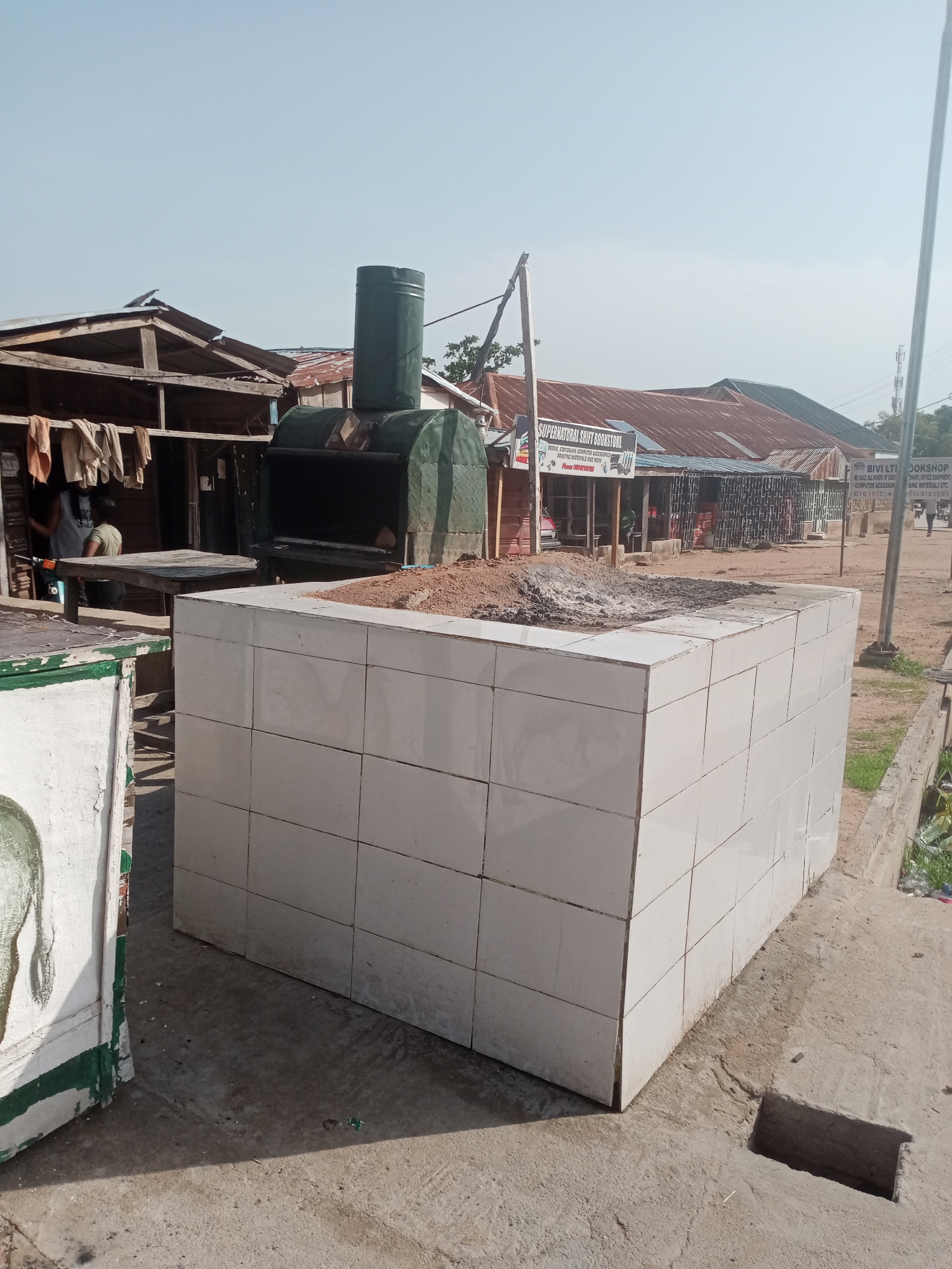 The image shows an outdoor scene in Jalingo, Nigeria. In the foreground, a large, tiled structure, possibly a monument or a part of a building, is visible. A pile of dirt is on top of this structure. Behind it, a large, dark green metal oven with a chimney stands. The oven appears to be industrial or for cooking. To the left of the oven, there is a wooden stall or shop with clothes hanging on display. In the background, several other buildings with corrugated iron roofs line a street. Signs for "SUPERNATURAL SHIFT BOOKSTORE" and "BIVI LTI BOOKSHOP" are visible on some of the buildings, indicating a commercial area. A few people are present, and the overall environment suggests a busy, possibly market-like, area under a clear sky.