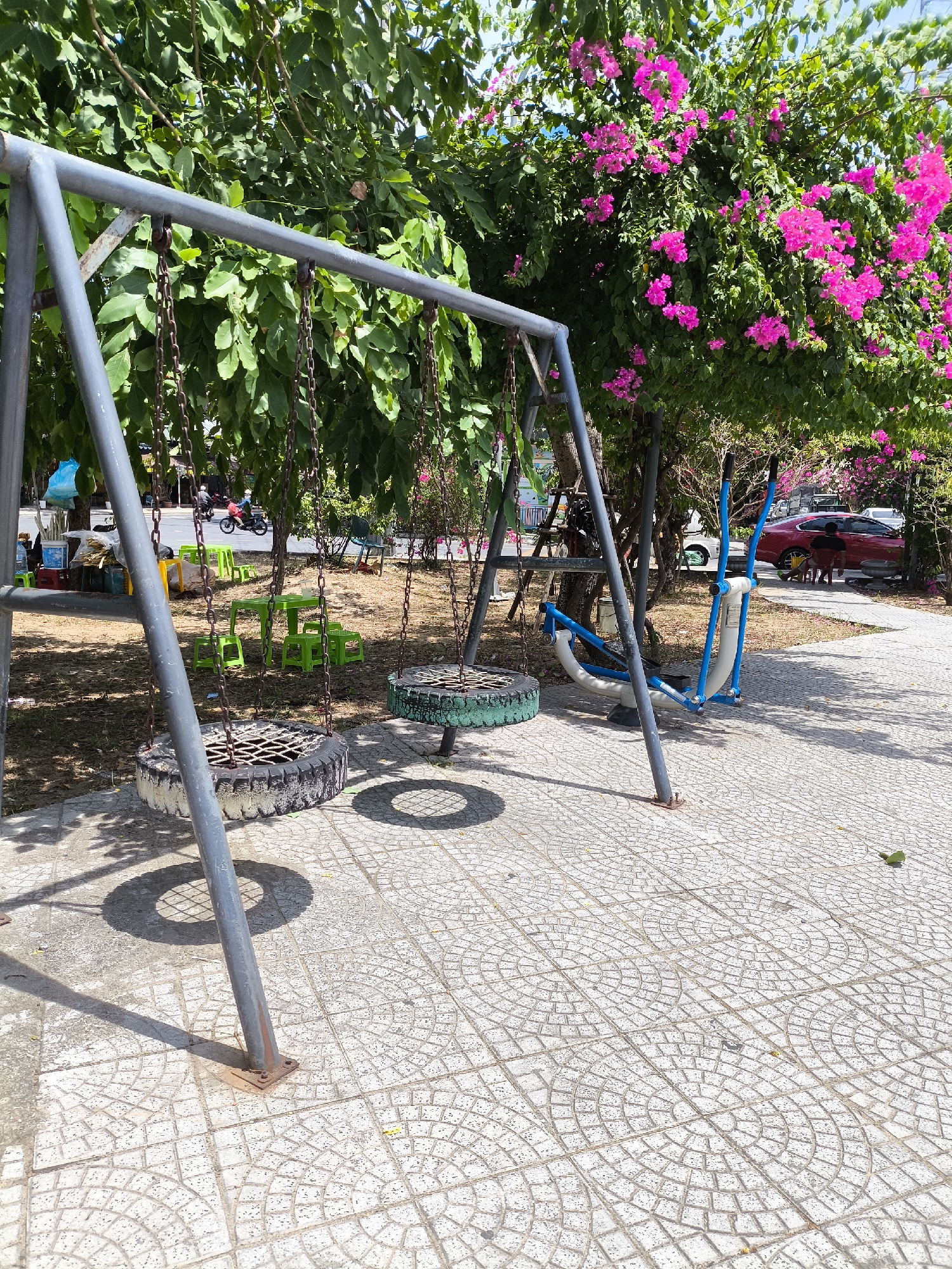 The image shows an outdoor public space in Da Nang, Vietnam, featuring a swing set with two tire swings and an elliptical exercise machine. The ground is paved with patterned tiles. Green plastic tables and chairs are visible in the background, along with trees and bright pink bougainvillea flowers. A street with a motorcycle and a car can be seen beyond the playground area. The scene appears to be set in a park or a recreational area in a city.