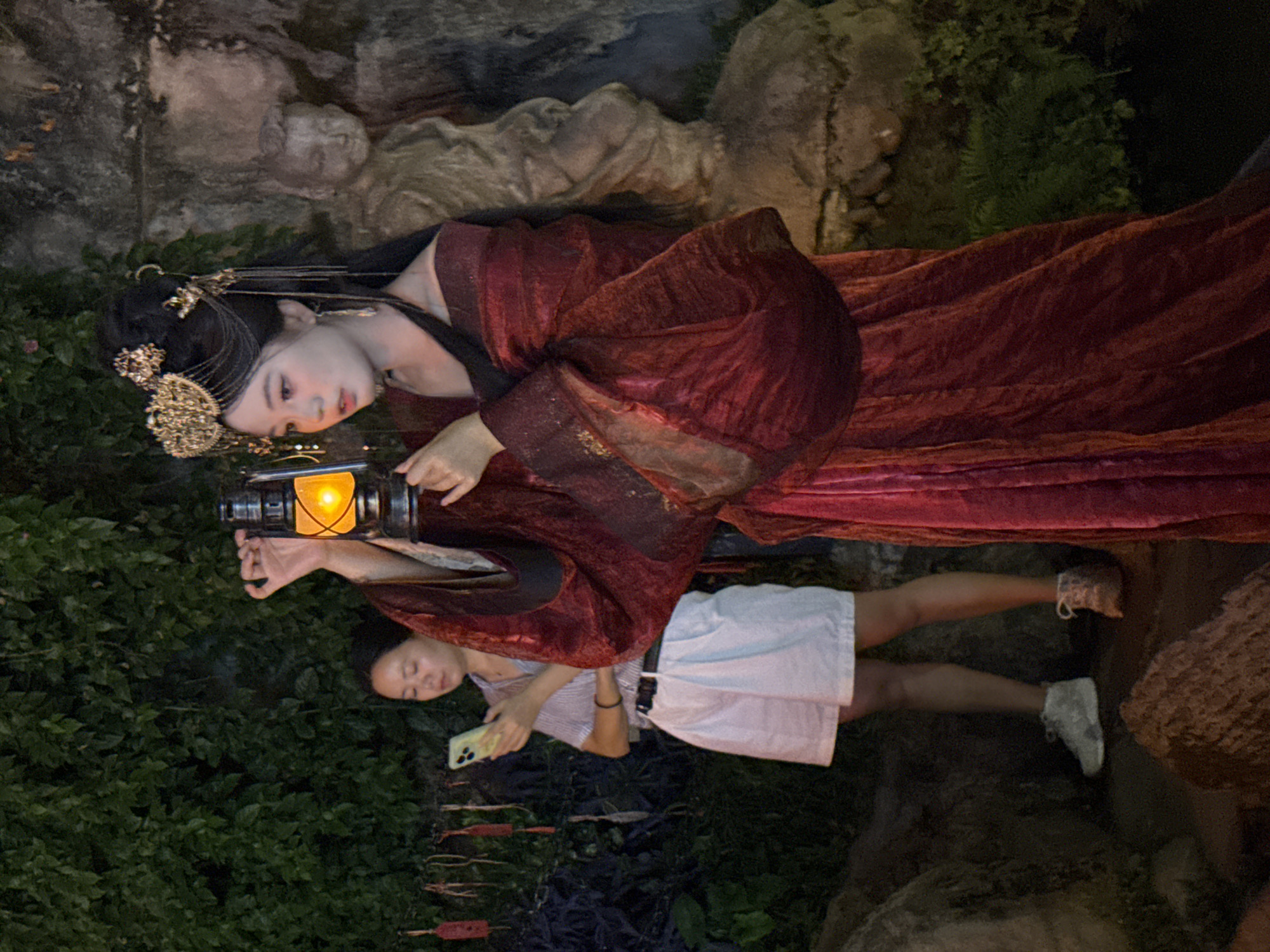 A young woman in a traditional red Chinese dress and elaborate gold headdress holds an illuminated lantern aloft. Her makeup is dramatic, with prominent eyeliner and blush. Behind her, slightly out of focus, a second young woman in a striped shirt and white shorts is looking at her phone. The background is a dark, naturalistic setting with lush green foliage and a stone wall or rock formation. A small, carved stone figure is visible behind the woman with the lantern. The scene appears to be in a garden or park-like setting, possibly at dusk or night, given the artificial light of the lantern and the dim surroundings. The location is identified as Yuzhong District, China.
