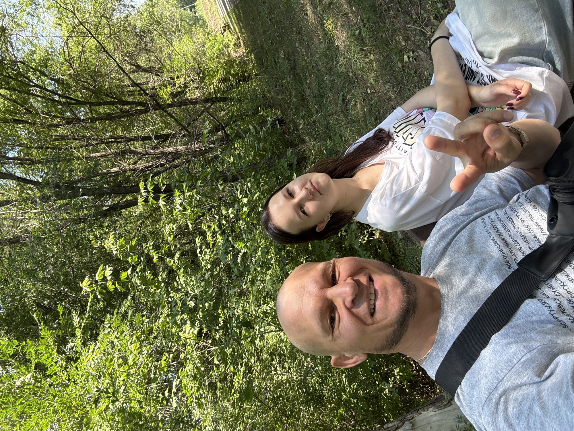 The image shows two individuals, a bald man with a beard and a woman with long dark hair, sitting on a park bench outdoors. The man is in the foreground, smiling and making a peace sign with his right hand, which is adorned with a bracelet. He is wearing a grey t-shirt with text printed on it and a black strap across his chest. The woman is to his right, wearing a white t-shirt with a graphic design and grey pants. She is looking towards the camera with a slight smile. They are positioned in front of a dense background of green trees and foliage, indicative of a park or wooded area. The ground around them is covered in grass and some fallen leaves. The lighting suggests it is daytime, possibly a sunny afternoon. No specific landmarks or identifying features of Oskemen, Kazakhstan are visible in the image.