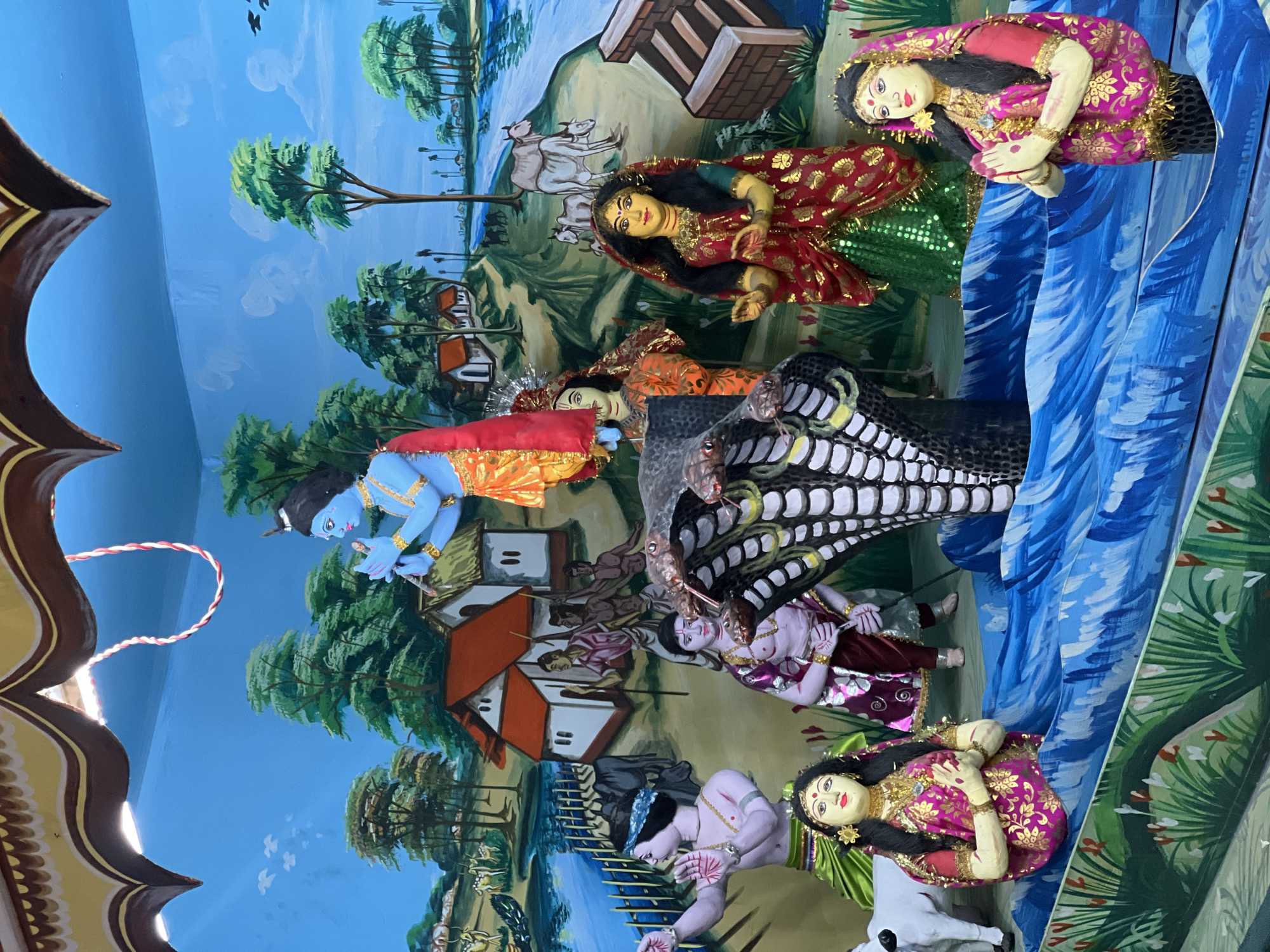The image depicts a three-dimensional tableau of Krishna subduing Kaliya, the venomous serpent king, set against a painted backdrop. The scene likely portrays an event from Hindu mythology, where Krishna, as a child, jumped into the Yamuna River to defeat Kaliya, who had poisoned the waters.
The scene features:
* **Krishna:** Depicted with blue skin, a common depiction of the deity. He stands on the head of Kaliya.
* **Kaliya:** Represented as a large, black serpent with numerous heads.
* **Gopis:** The women of Vrindavan. The scene features several Gopis on either side of the serpent king.
* **Painted Backdrop:** A blue sky with depictions of houses, trees, cows, and people.
The location may very well be in Varanasi, India. The image shows what is commonly called a Jhaki (a decorative display, a tableau), often made for religious purposes. It is made of paint, clay, and plaster. The tableau has a religious and cultural context, which is reflected in the elements of the scene.