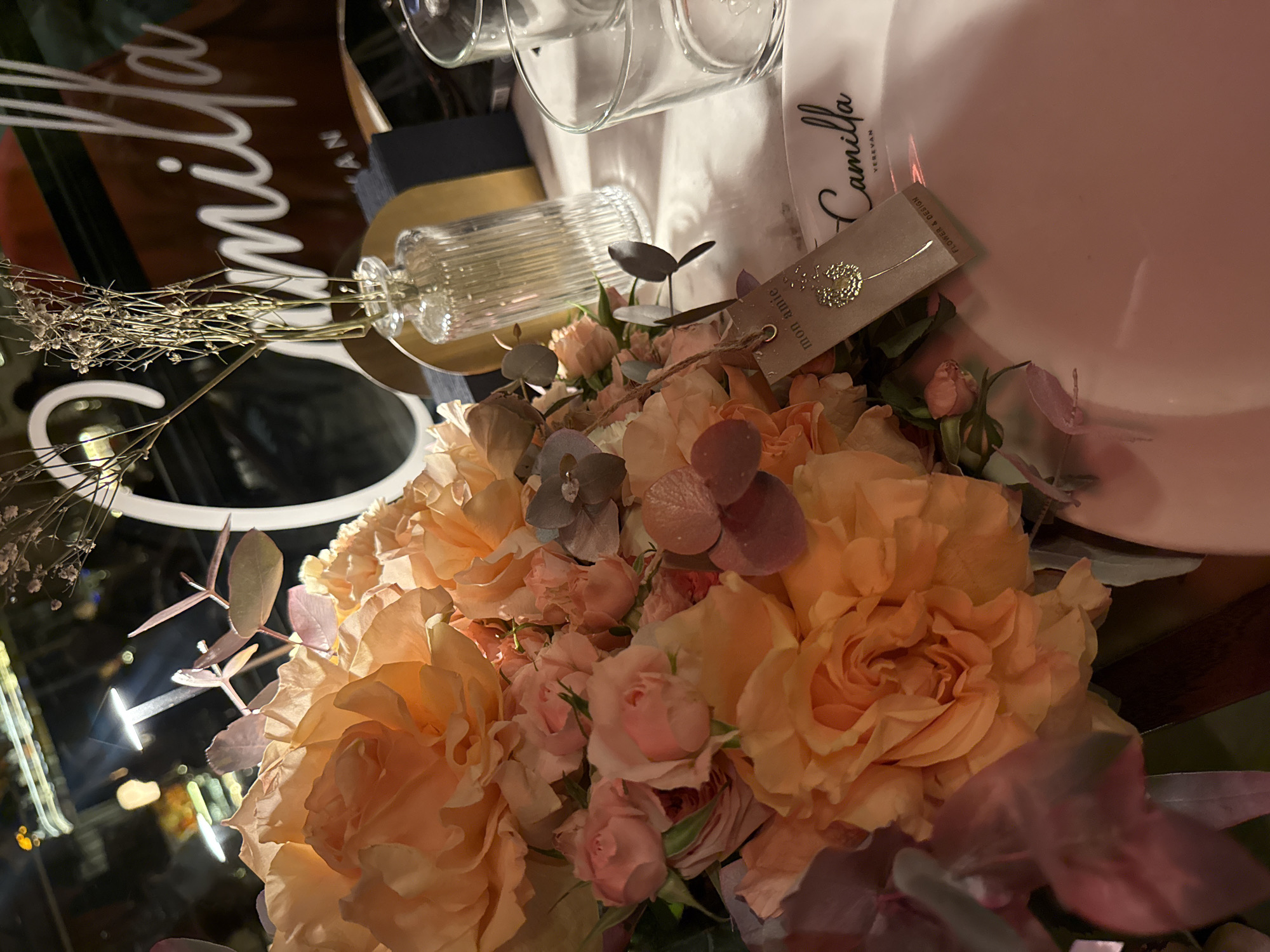 The image depicts a table setting, likely in a restaurant, featuring a floral centerpiece. The arrangement is composed of peach and pink roses with sprigs of dried flowers and purple foliage accents. Next to the flowers is a small, clear glass vase on a gold stand. Behind the vase and flowers are several wine glasses, a white cloth with the inscription "Camilla Yerevan," and a plate with the same writing. The tag in the flowers says "mon amie" which also bears the name of a flower company. The table appears to be reflective, possibly made of glass. The location of the scene is Yerevan, Armenia, as indicated by the inscription.