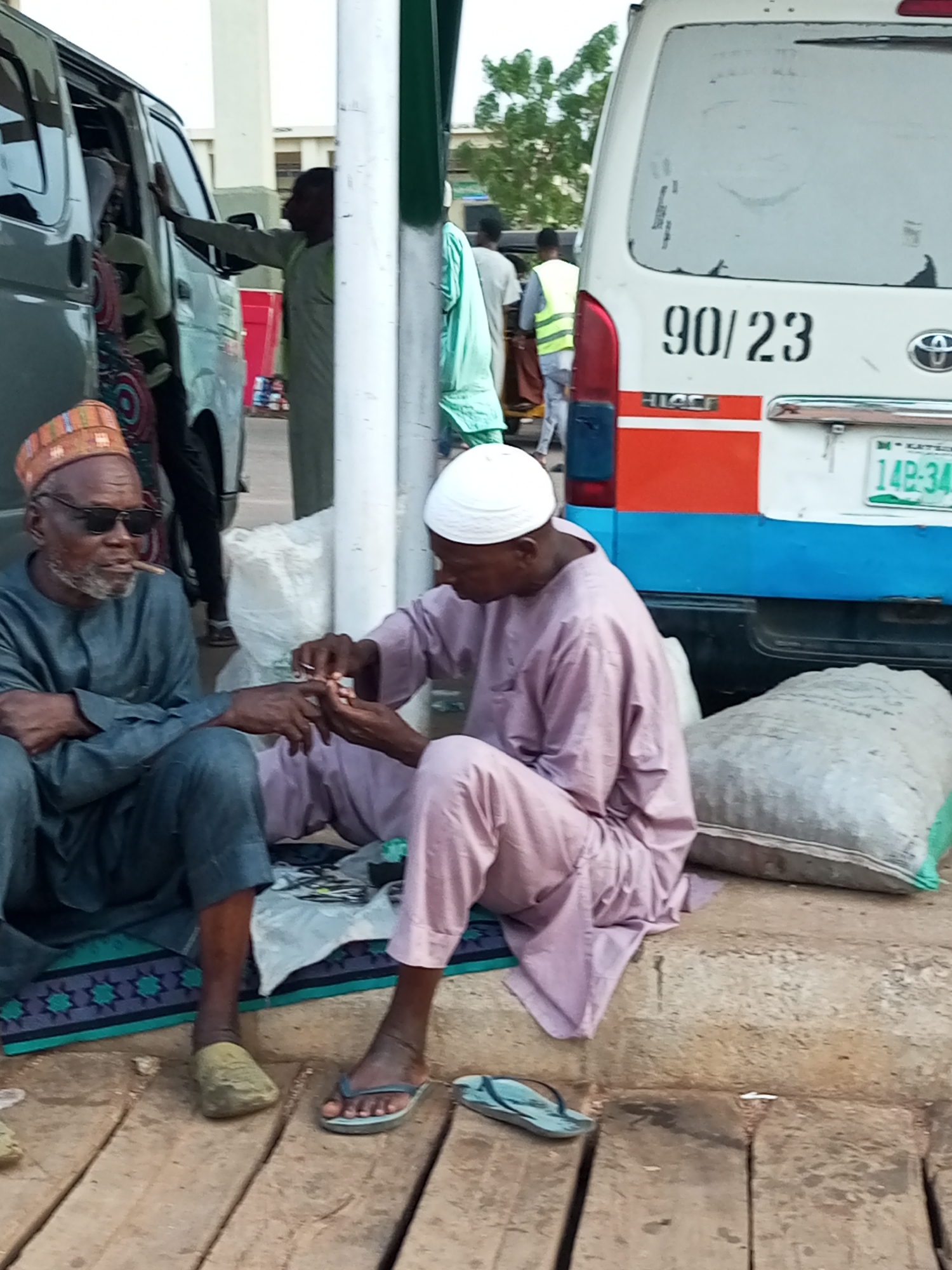 In Gombe, Nigeria, two men are seated on a raised concrete curb. The man on the left, wearing a dark grey traditional outfit and a patterned cap, has sunglasses and is smoking a cigar. The man on the right, dressed in a pale purple traditional outfit and a white cap, is attending to the first man's hands, appearing to be trimming his fingernails with a small tool. A large, light-colored sack is positioned behind them. In the background, a white van with "90/23" painted on its rear and a Nigerian license plate is visible, along with a silver minivan and several people. The ground is paved with wooden planks.