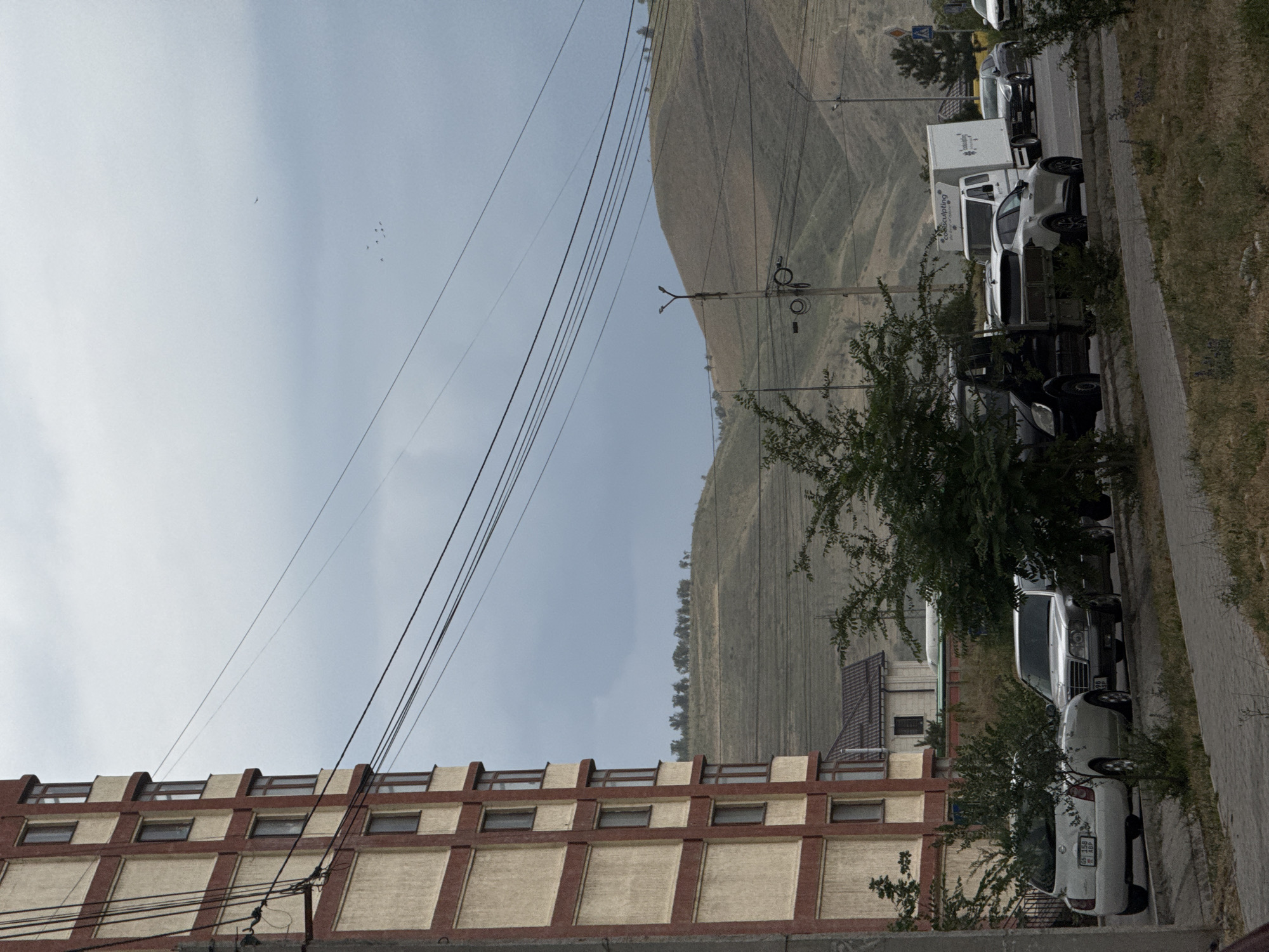 In a street scene in an unknown city, a multi-story building with a beige facade and red trim stands on the left. Power lines crisscross the sky above the building and the street. Beyond the building, rolling hills with sparse vegetation are visible under a cloudy sky. The street in the foreground has several parked cars, including a white hatchback and a silver sedan. A white delivery truck and other vehicles are also present on the road. Green trees and bushes line the sidewalk and the edge of the road, with some obstructing the view of the cars. The overall scene depicts a residential or commercial street with a backdrop of natural hills.