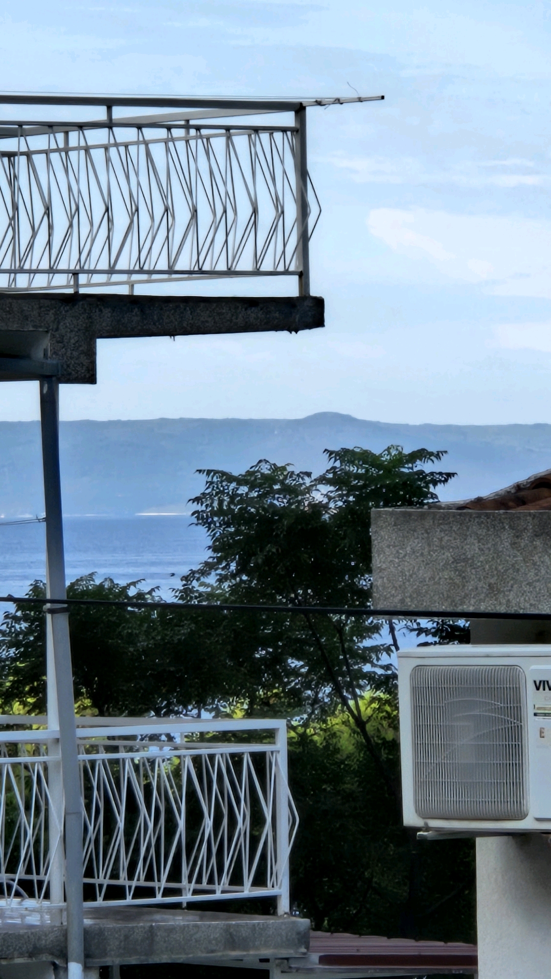 The image captures a view from a balcony or terrace in Općina Tučepi, Croatia. The foreground features a modern metal railing with a geometric pattern, likely made of steel. Below this railing, a concrete structure is visible. Further down, another white metal railing with a similar crisscross design is present, suggesting multiple levels or adjacent outdoor spaces.
In the mid-ground, lush green trees with dense foliage create a natural barrier. Behind the trees, a body of water, presumably the Adriatic Sea, stretches out, reflecting the sky. In the distance, a range of hazy, blue mountains forms the horizon, characteristic of the Dalmatian coast.
To the right of the frame, an outdoor air conditioning unit is mounted on a beige concrete wall. The unit is white with a visible grill. The sky above is a pale blue with scattered white clouds, indicating a partly cloudy day. The overall scene conveys a sense of a residential or hotel setting with a scenic view of the coast and surrounding landscape. There are no people or animals visible in the image.