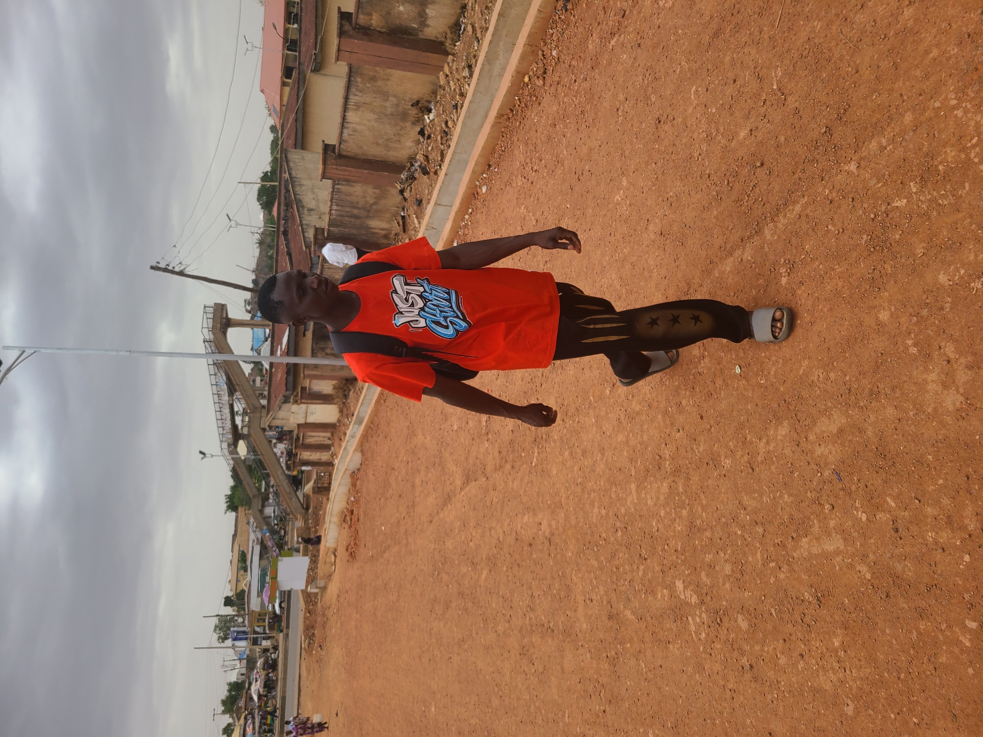 A young man wearing a red t-shirt with blue lettering, black patterned leggings, and slides stands on a dusty red-brown surface. He has a black backpack on. In the background is a cityscape under a cloudy sky, with visible buildings, a pedestrian bridge, power lines, and a street with activity. The location is likely in the city of Jos, Nigeria, suggested by the style of the environment and urban architecture.