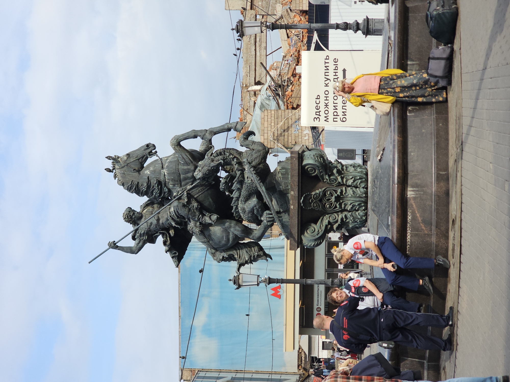 The image captures a statue of Saint George slaying a dragon, a prominent monument located in Moscow, Russia. The statue is positioned in front of a backdrop that includes a large blue banner with a sky motif, a Moscow Metro station entrance with the iconic red 'M' logo, and a section of a building undergoing demolition, evident by scattered bricks and debris. Several people are present in the scene. In the foreground, a man in a dark blue uniform with red insignia is seen talking to two women, also in uniforms with red patches. To the right, a woman in a yellow jacket and a patterned skirt stands near a sign with Russian text, a large tote bag on her shoulder and two pieces of luggage on the ground. The overall environment suggests an urban setting, possibly a public square or transit hub, with elements of both active city life and ongoing construction or renovation.