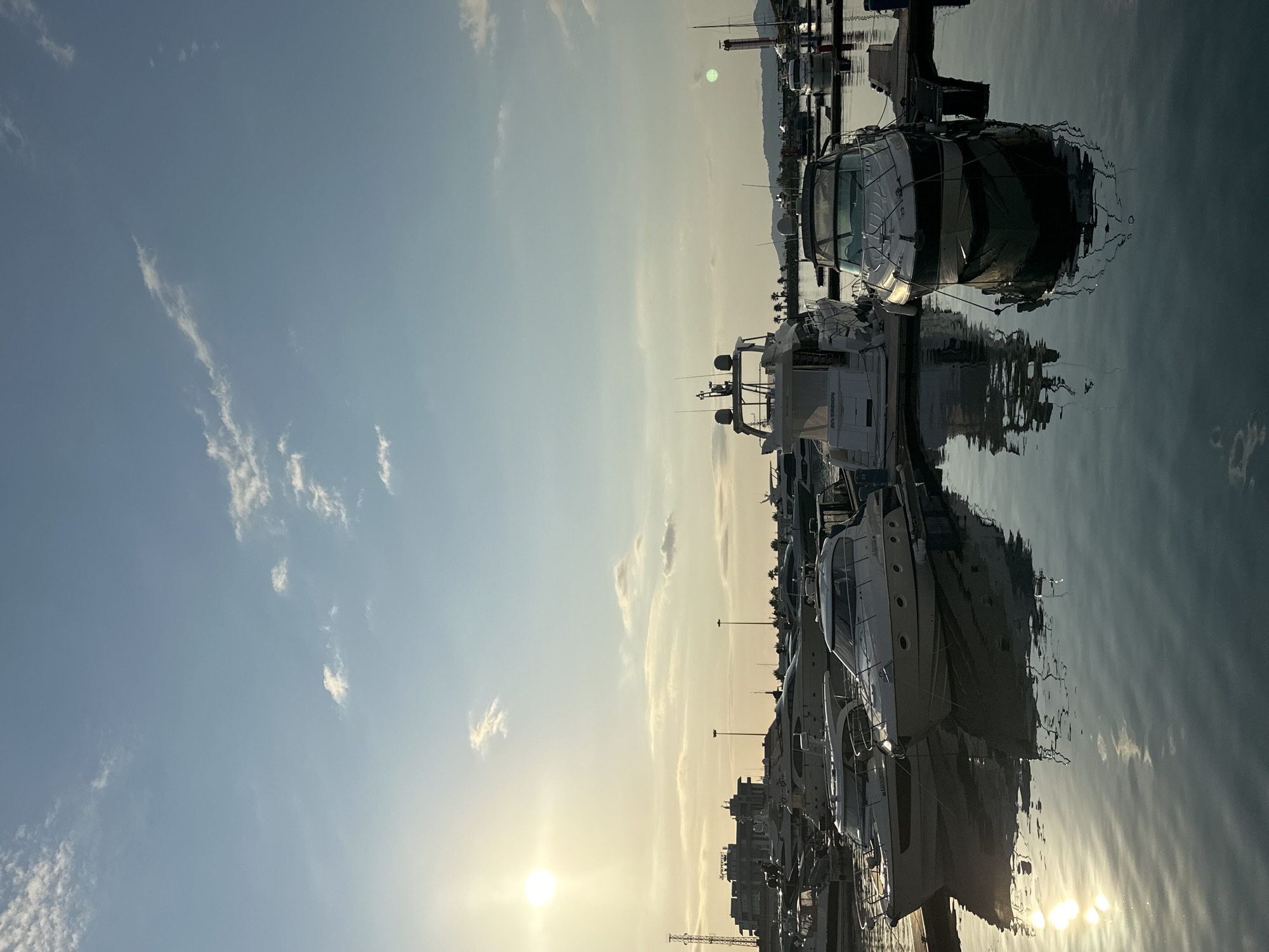 In Sochi, Russia, a marina is depicted with several yachts docked under a bright, hazy sun. The sky is a pale blue with scattered wisps of white clouds. The sun, positioned in the upper left quadrant, casts a warm, golden light across the scene, reflecting on the calm water.
The foreground features several white motor yachts, two of which are prominently docked side-by-side. These yachts have sleek designs, with visible cabins, railings, and antennae. Their reflections are clearly visible on the glassy surface of the water.
In the middle ground, more yachts of various sizes are moored. Beyond the docked vessels, a cityscape emerges with modern buildings and tall lampposts. Further in the distance, a line of palm trees and hills can be seen, suggesting a coastal or near-coastal location. The overall atmosphere is serene and tranquil, characteristic of a peaceful afternoon in a marina. There are no people visible in the image.