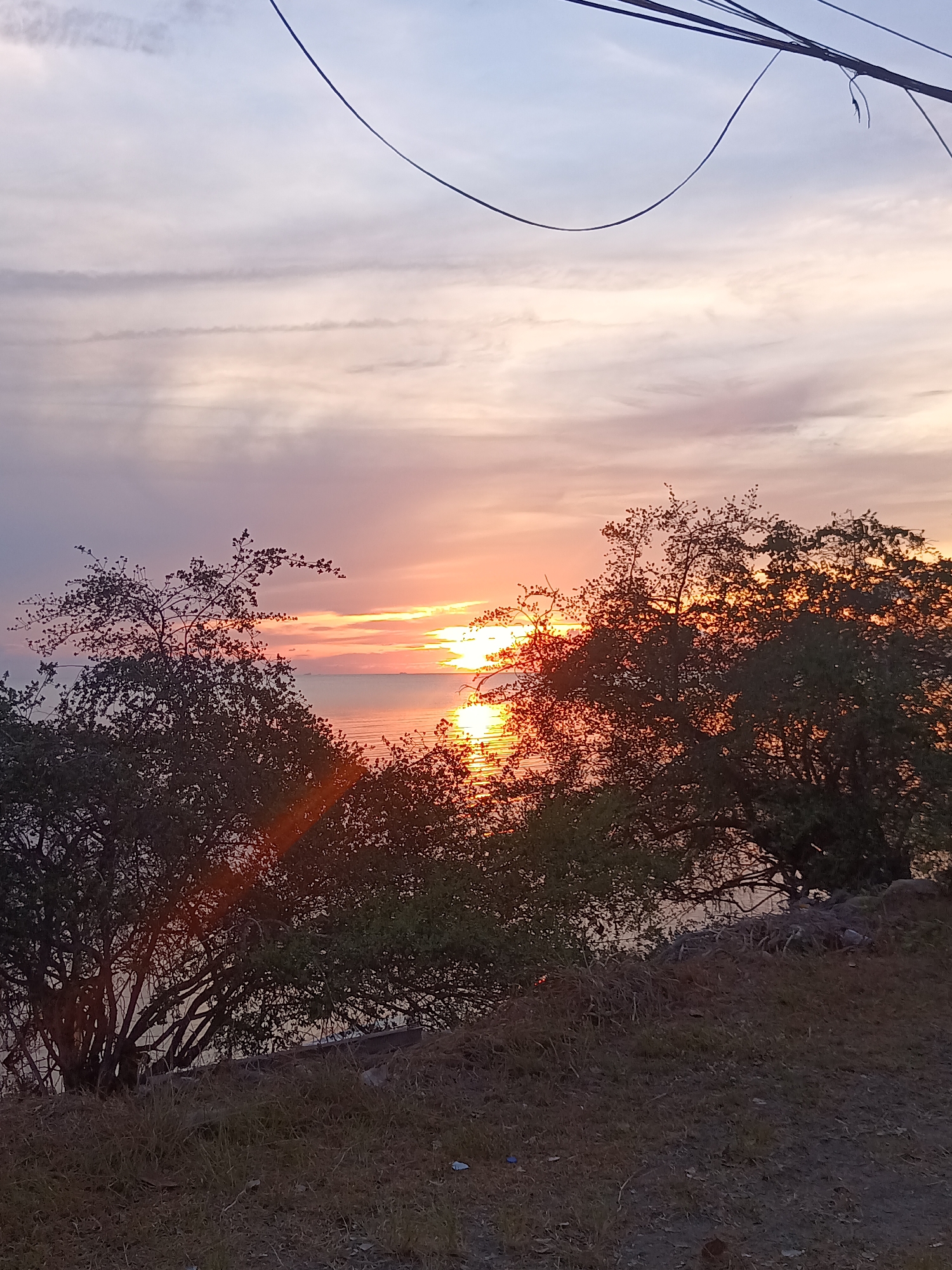 This image depicts a sunset over water, viewed from an elevated position. The location is Baan Sri Thanu, Thailand.
The sky shows a gradient of colors, from blue at the top to orange and pink near the horizon. Wispy clouds are visible across the sky. In the center of the horizon, the sun is setting, creating a bright reflection on the water's surface. Silhouetted trees frame the view on both sides. The foreground shows a grassy slope with vegetation. Overhead, there are some unidentifiable cables or wires.